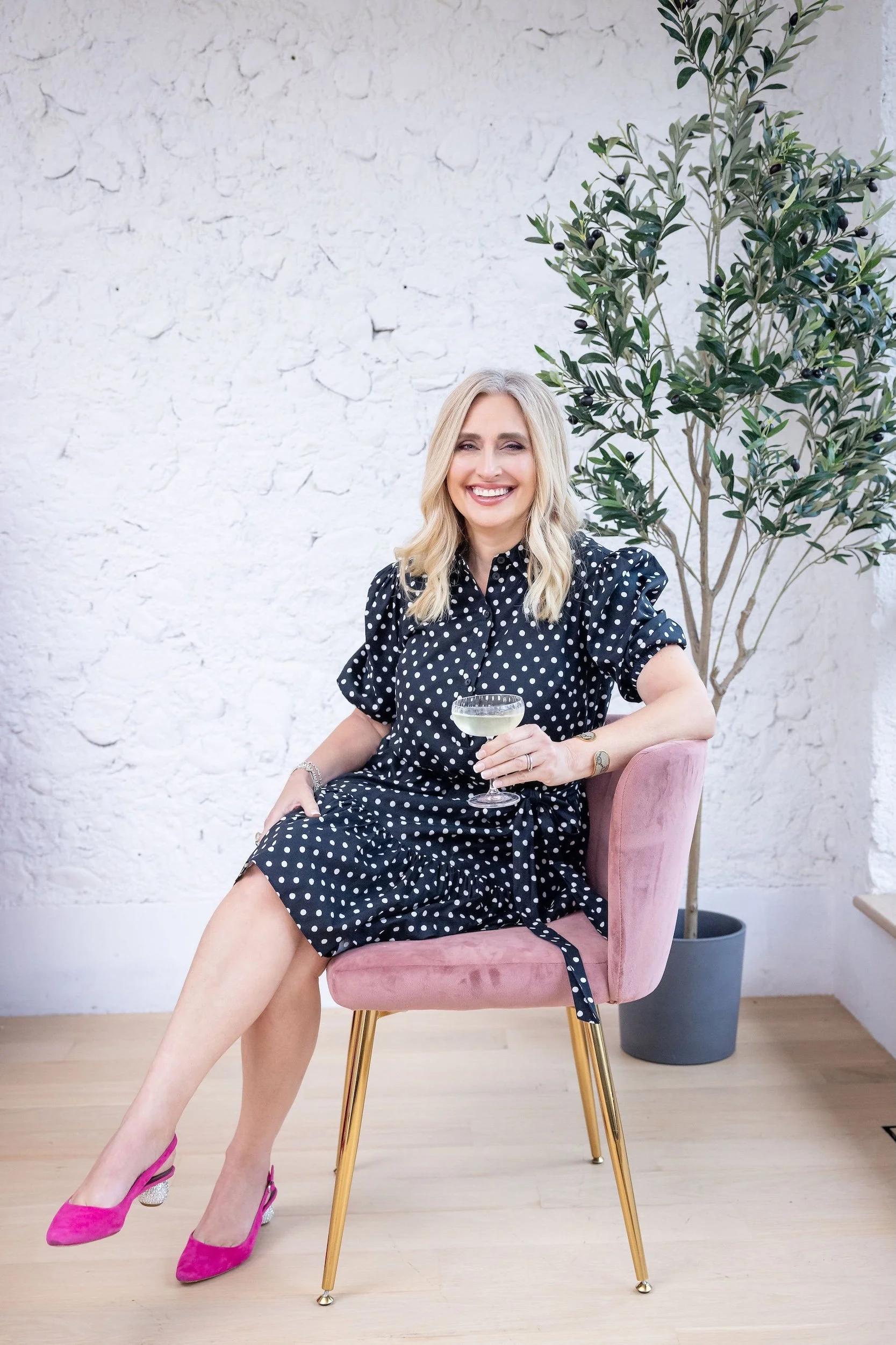 Lauren Brynjelsen wearing a black polka dot dress and pink high heels sitting on a pink velvet chair, holding a glass of white wine or champagne, smiling in front of a white textured wall with a tall green plant in a black pot.