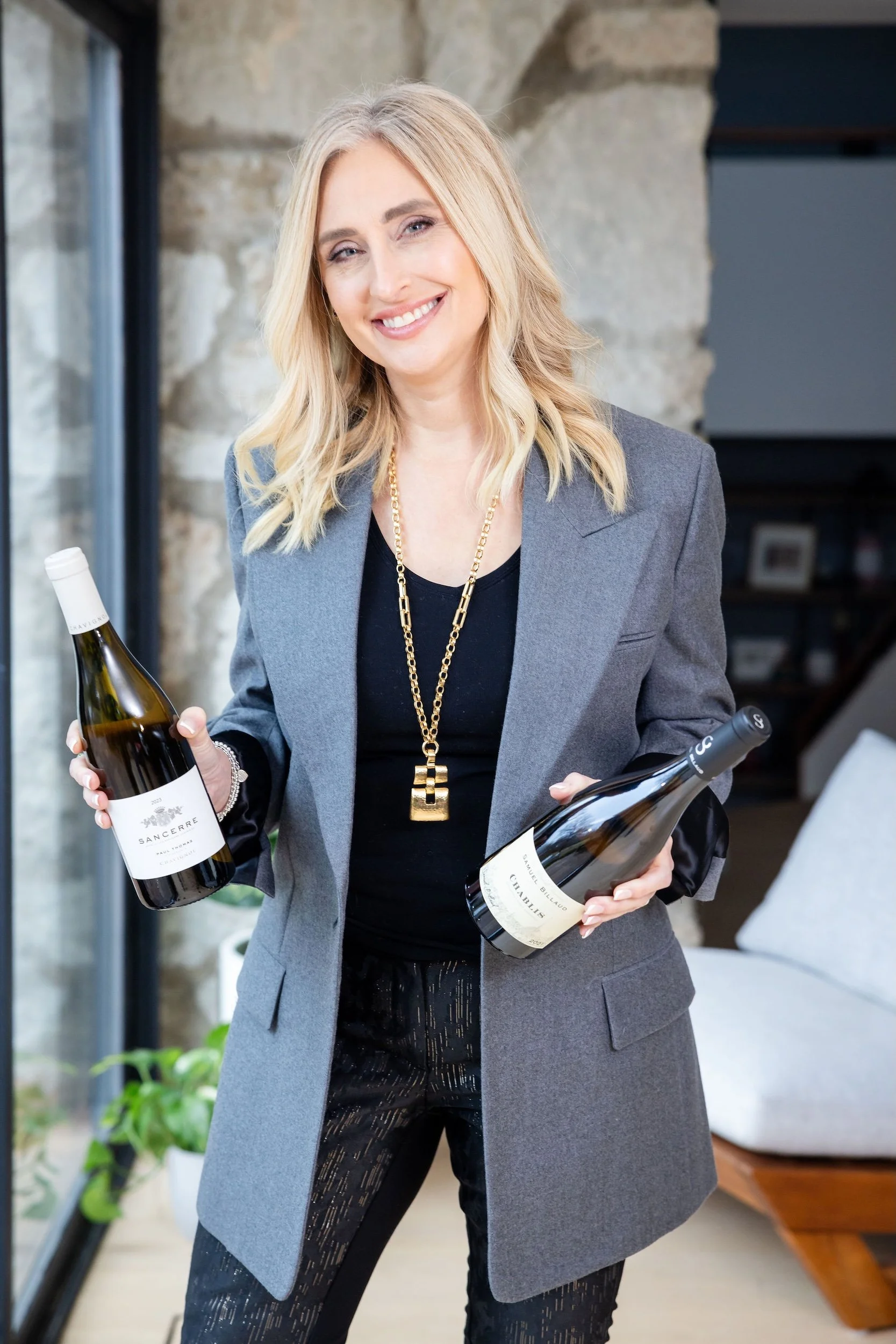 Lauren Brynjelsen with blonde hair smiling and holding two bottles of wine, one in each hand, indoors in front of a stone wall and a window.
