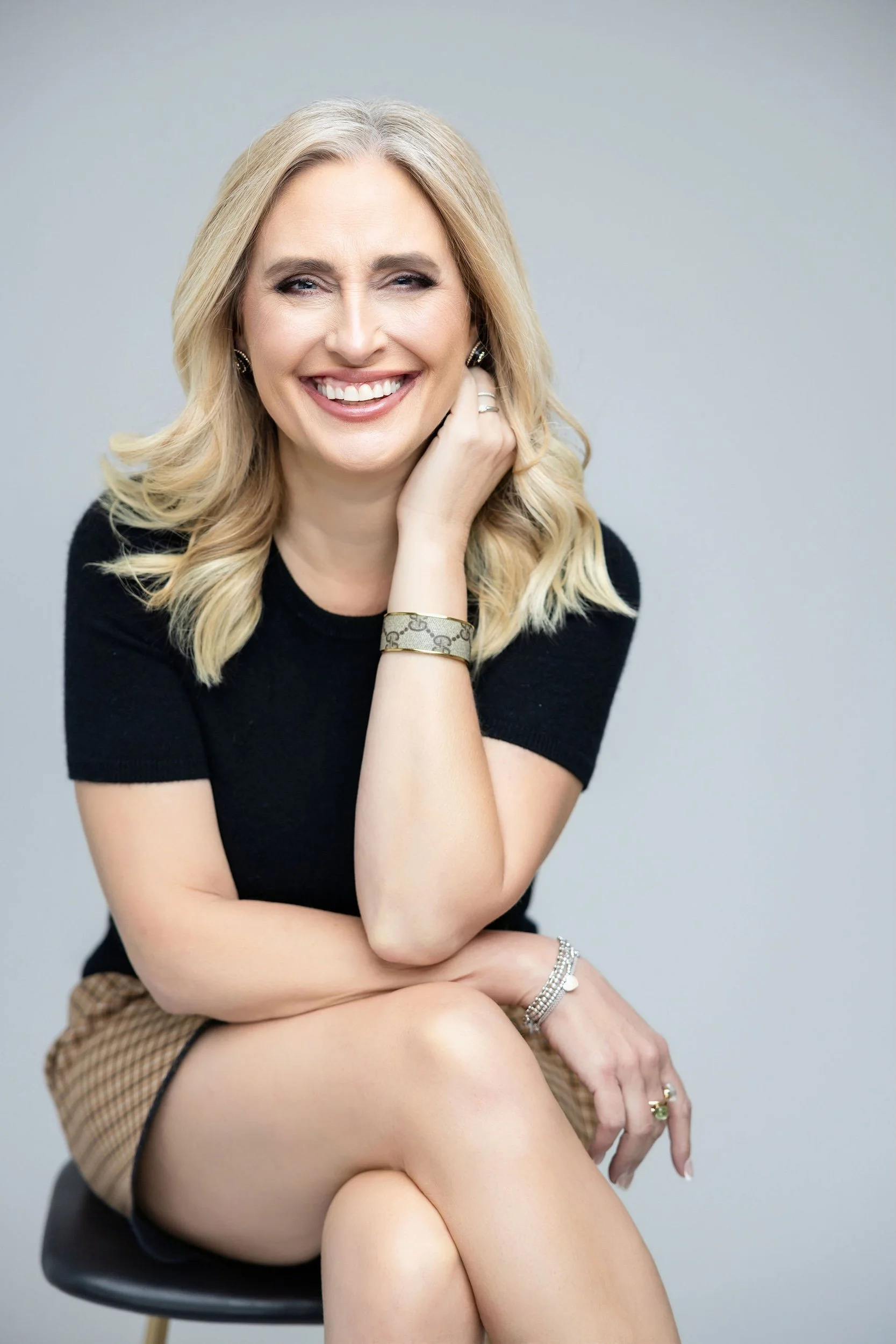 Lauren Brynjelsen wearing a black top and a tan skirt, sitting on a stool against a gray background.