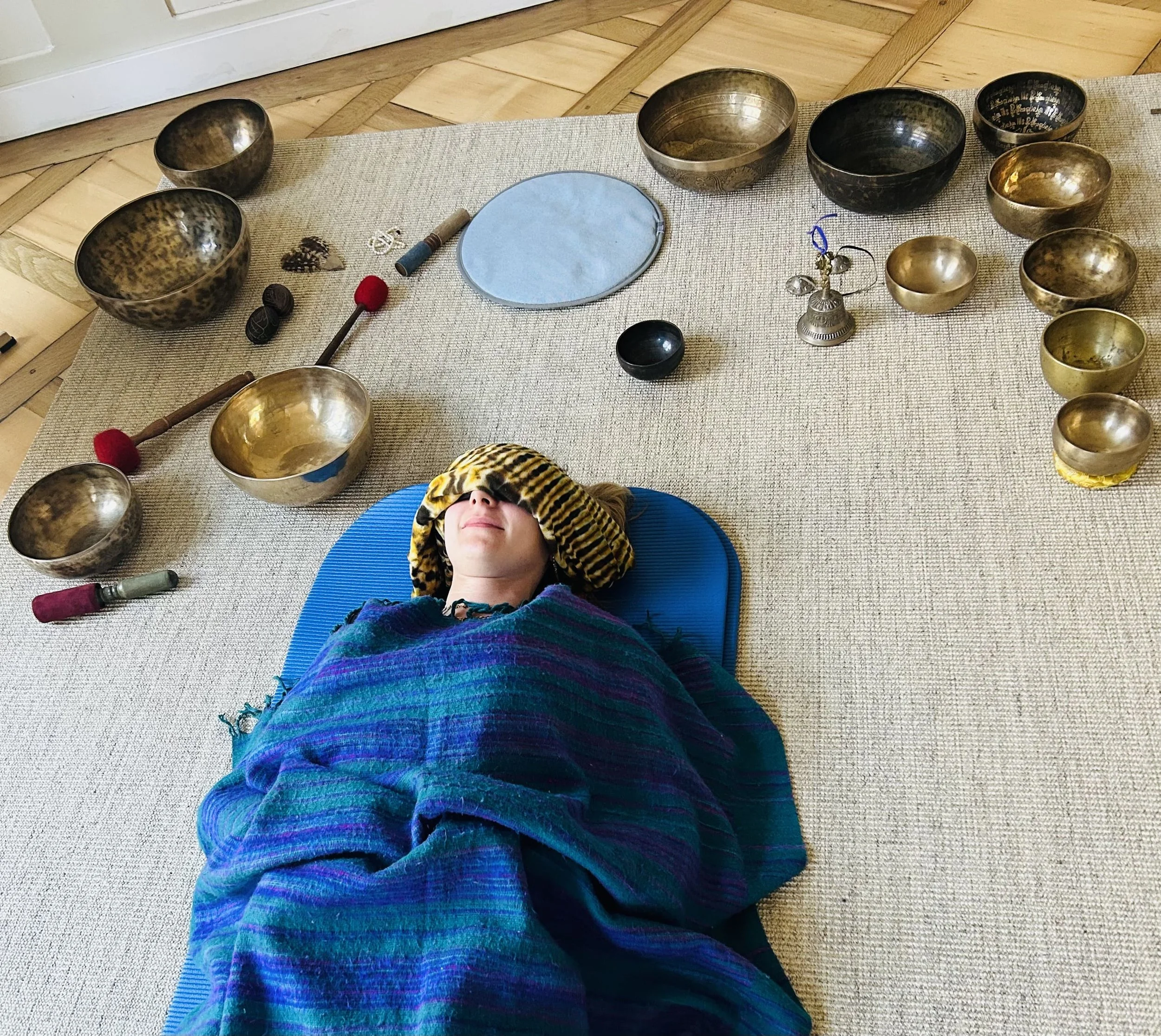 Eine Person liegt auf einem blauen Yogamatte, umgeben von Klangschalen, einer Gitarre, einer Tasche mit buntem Muster und sonstigen Gegenständen auf einem Teppichboden.