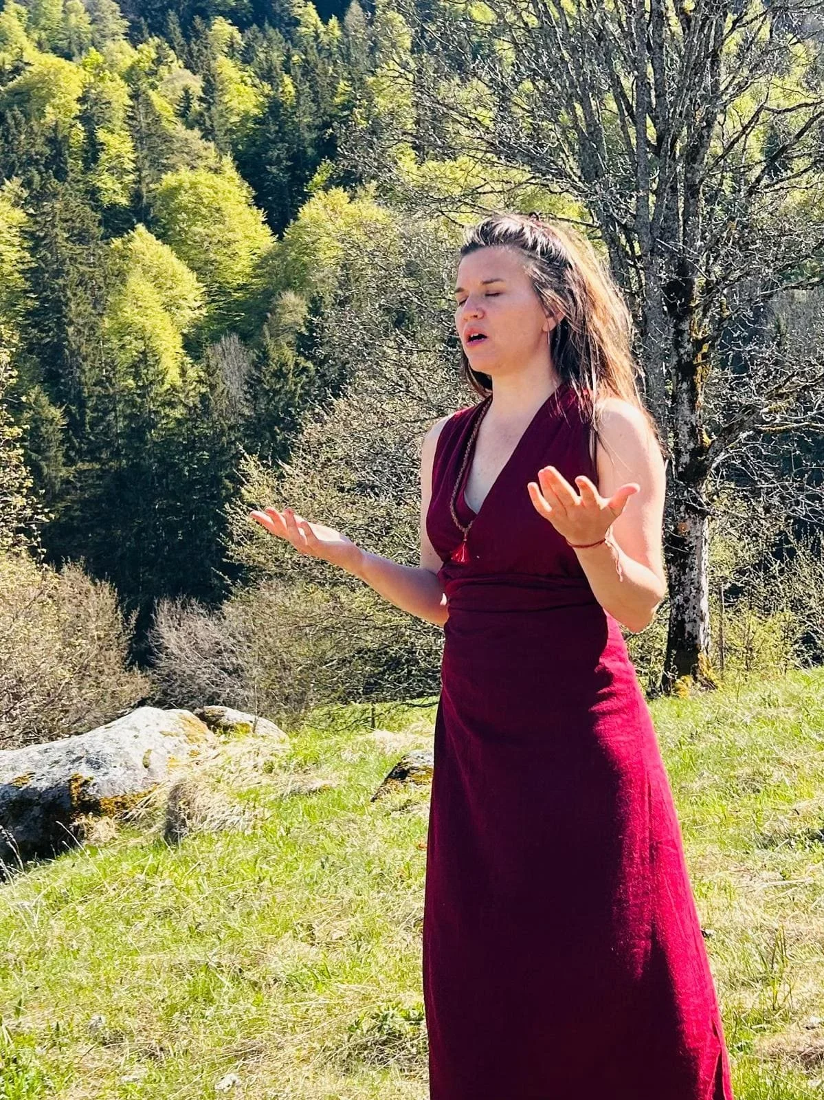 Eine Frau in einem langen weinroten Kleid steht draußen in der Natur, mit geschlossenen Augen und erhobenen Händen, als ob sie meditiert oder betet, umgeben von Bäumen und grünem Gras.