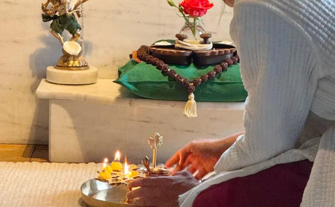 Person hält eine große Flöte mit Kerzen auf einem silbernen Tablett. Auf einem Tisch im Hintergrund stehen eine Statue, eine Vase mit roten Rosen, Gebetsketten und andere Gegenstände.