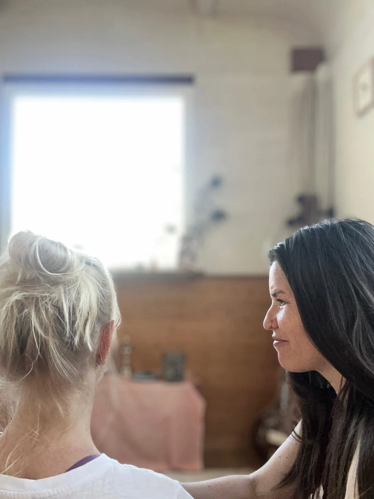 Zwei Frauen, eine ältere mit grauem Haar und eine jüngere mit schwarzen Haaren, sitzen sich im Wohnzimmer gegenüber, haben eine freundliche Interaktion, im Hintergrund ein Fenster, ein Couchtisch und ein Werkzeug.
