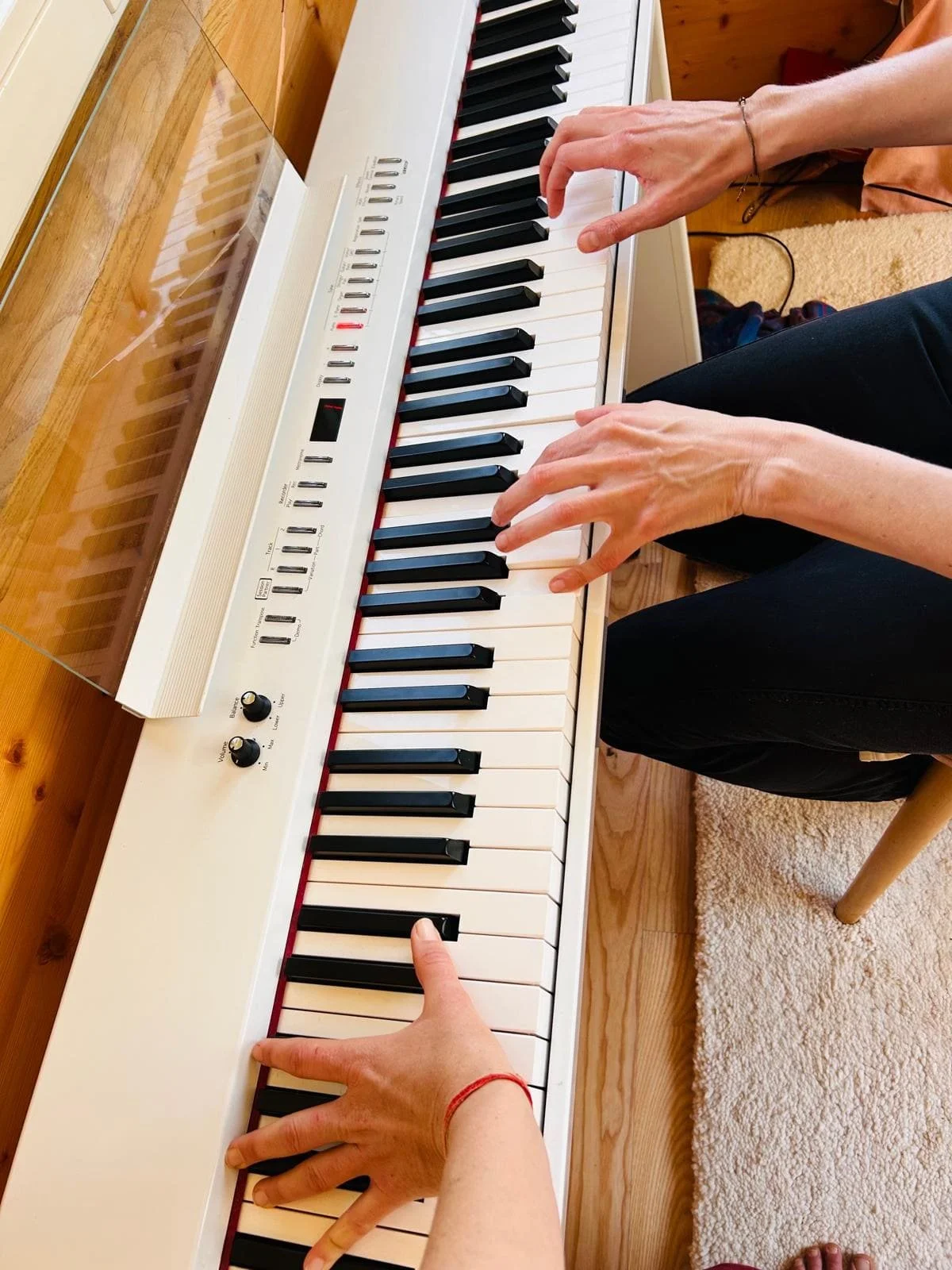 Zwei Personen spielen zusammen Klavier, die Hände sind auf den Tasten, im Hintergrund ein Holzfußboden und ein Teppich.