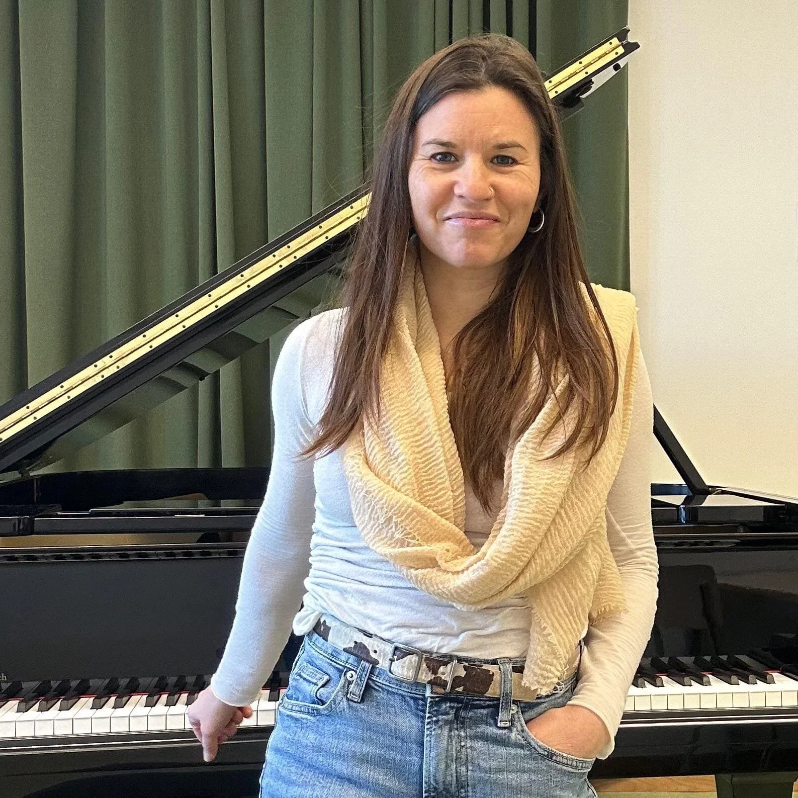 Eine Frau steht vor einem Klavier, trägt ein weißes Oberteil, eine gelbe Schal und eine Jeans, und lächelt leicht.