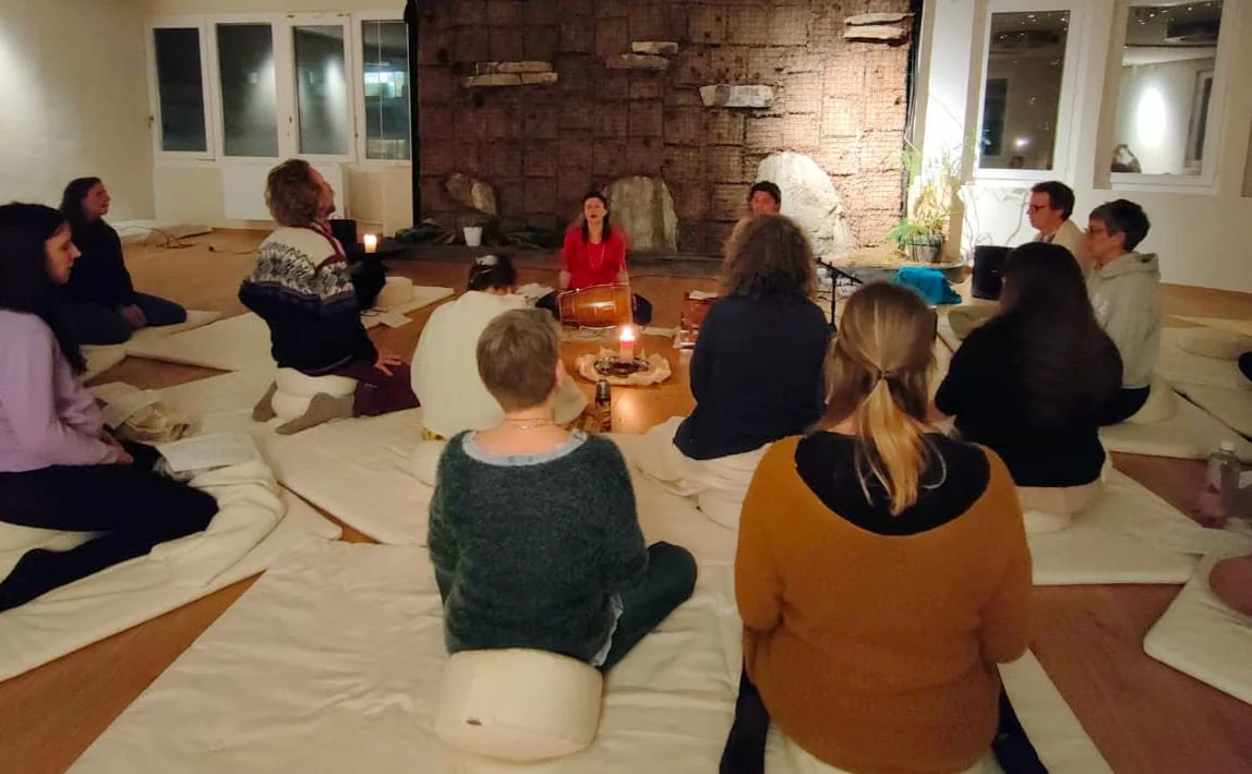 Menschen sitzen in einem Kreis auf Matratzen in einem Raum, einer Person singt oder spricht, im Hintergrund ist eine Wand mit Steinen und Pflanzen, ruhige Atmosphäre.