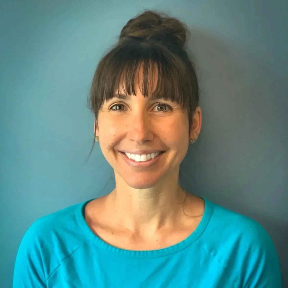 A woman with brown hair styled in a top bun, wearing a bright blue shirt, smiling, standing in front of a plain blue background.