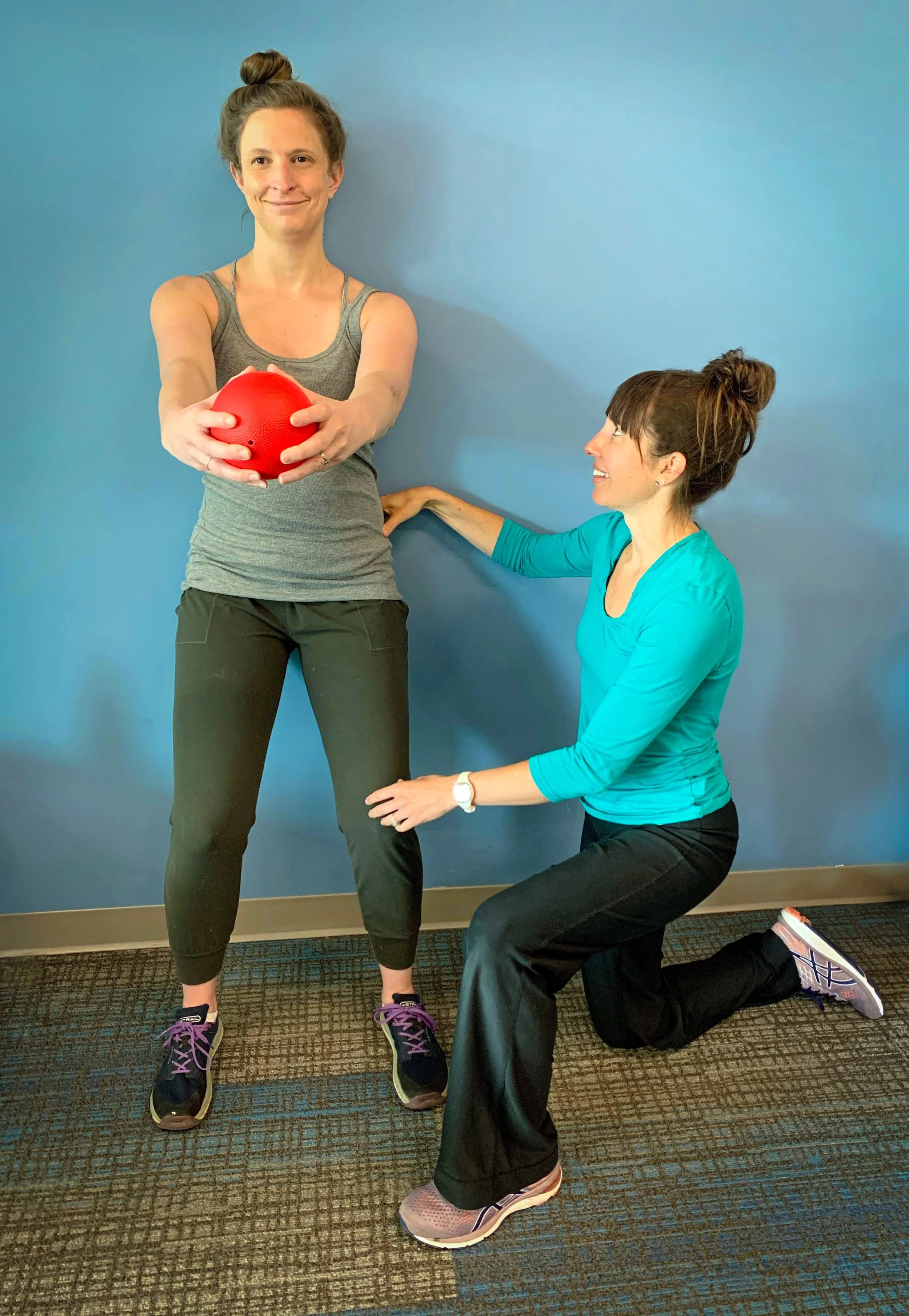 Woman standing and holding a red exercise ball with both hands, supervised by a trainer kneeling on one knee, supporting her knee, against a blue wall.