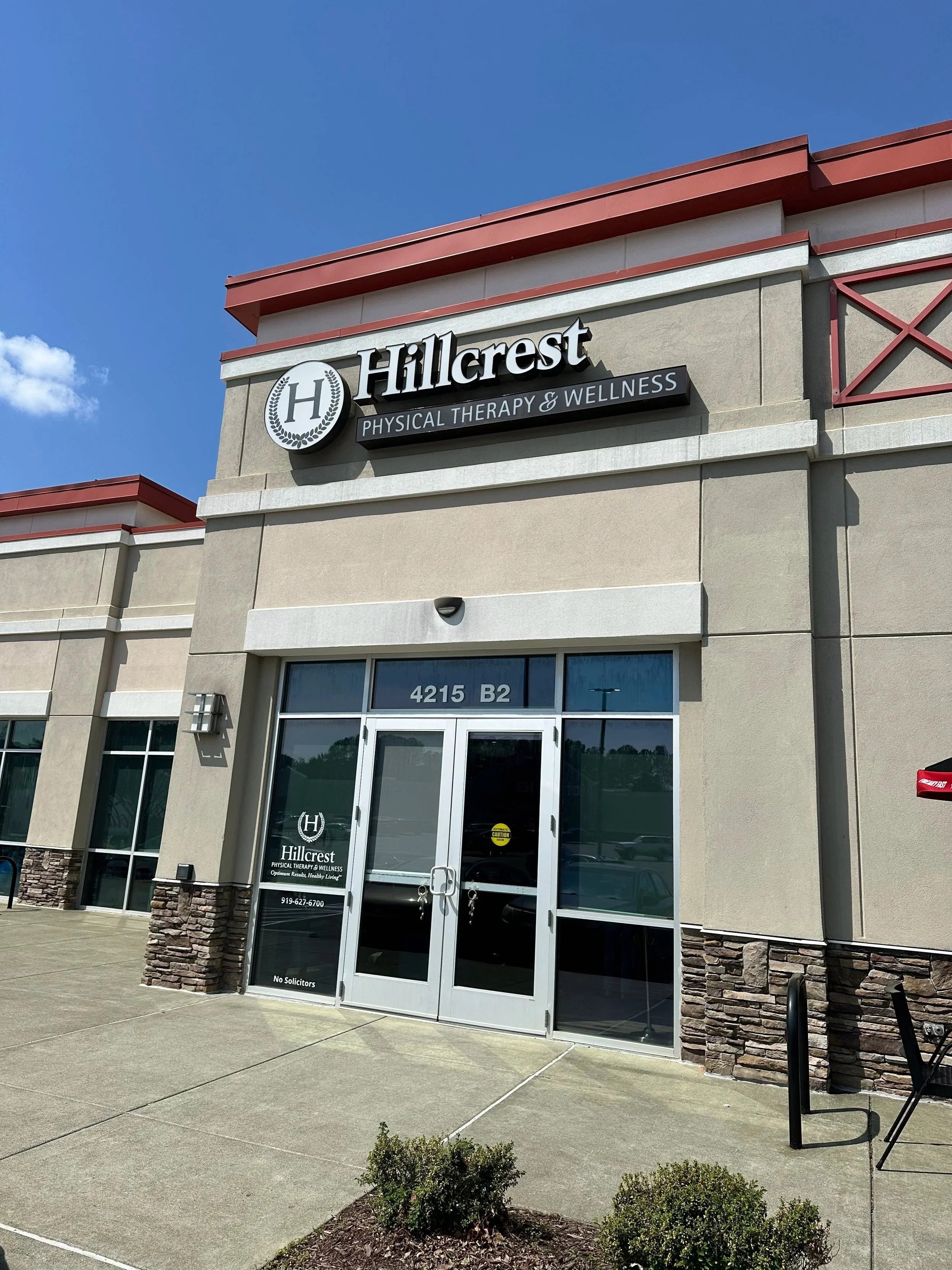 Exterior view of Hillcrest Physical Therapy & Wellness clinic with a beige building, glass doors, and signage, under a blue sky.