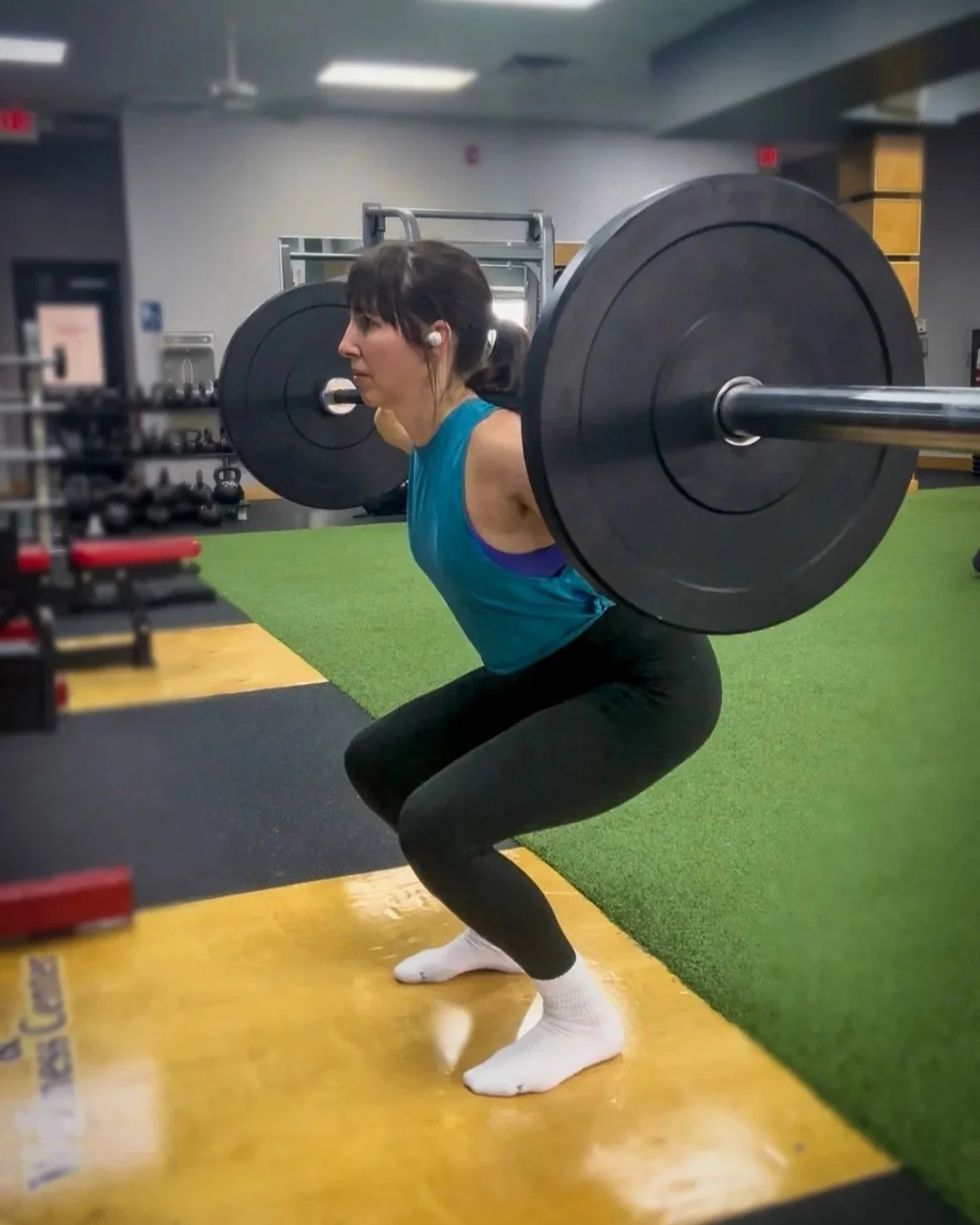 Woman performing a squat exercise with a barbell loaded with large black weight plates in a gym setting.