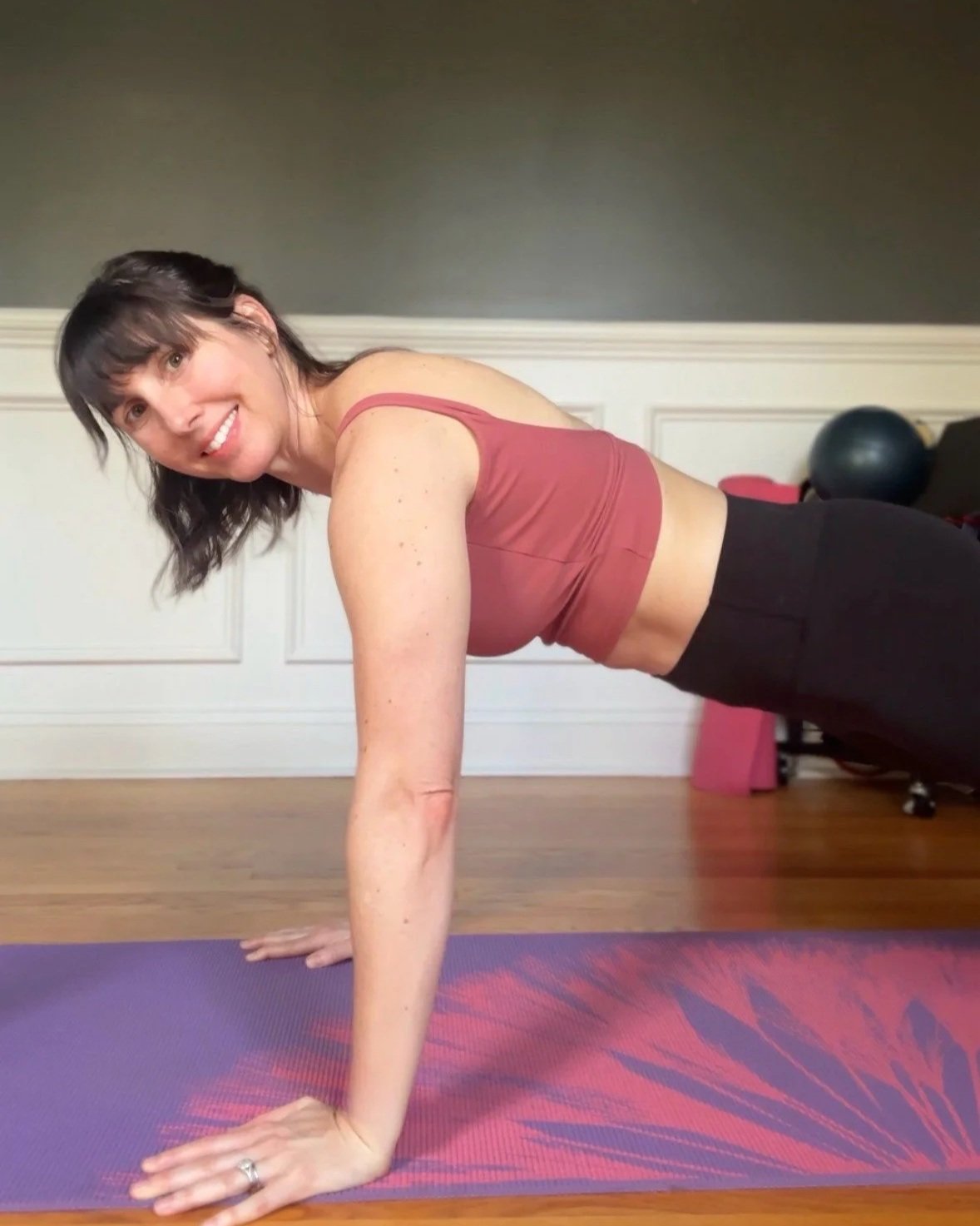 Woman in workout clothes smiling while doing a plank exercise on purple yoga mat in a room with hardwood floor and exercise equipment in the background.