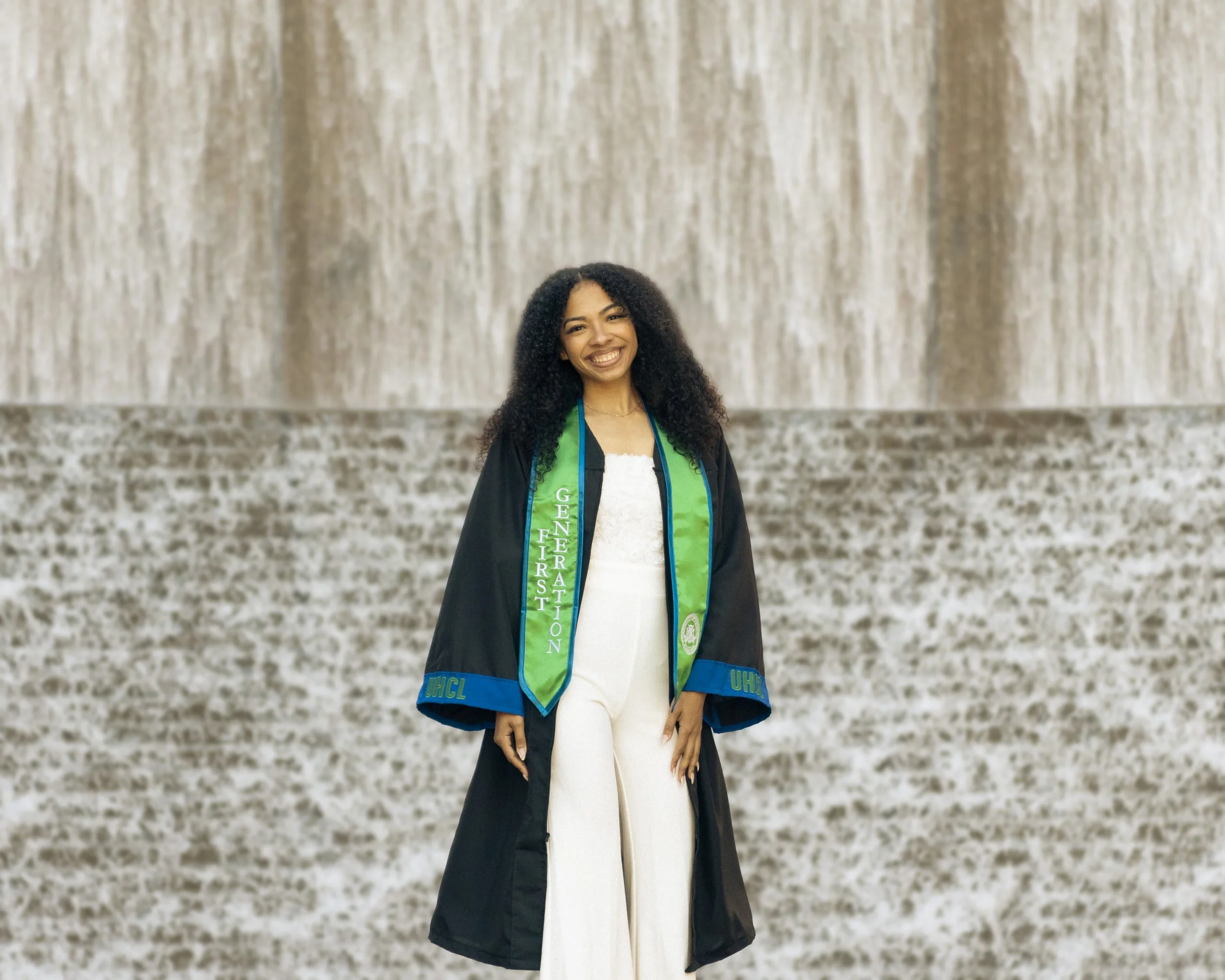 A young woman in a graduation gown and a green and blue stole standing in front of a waterfall.