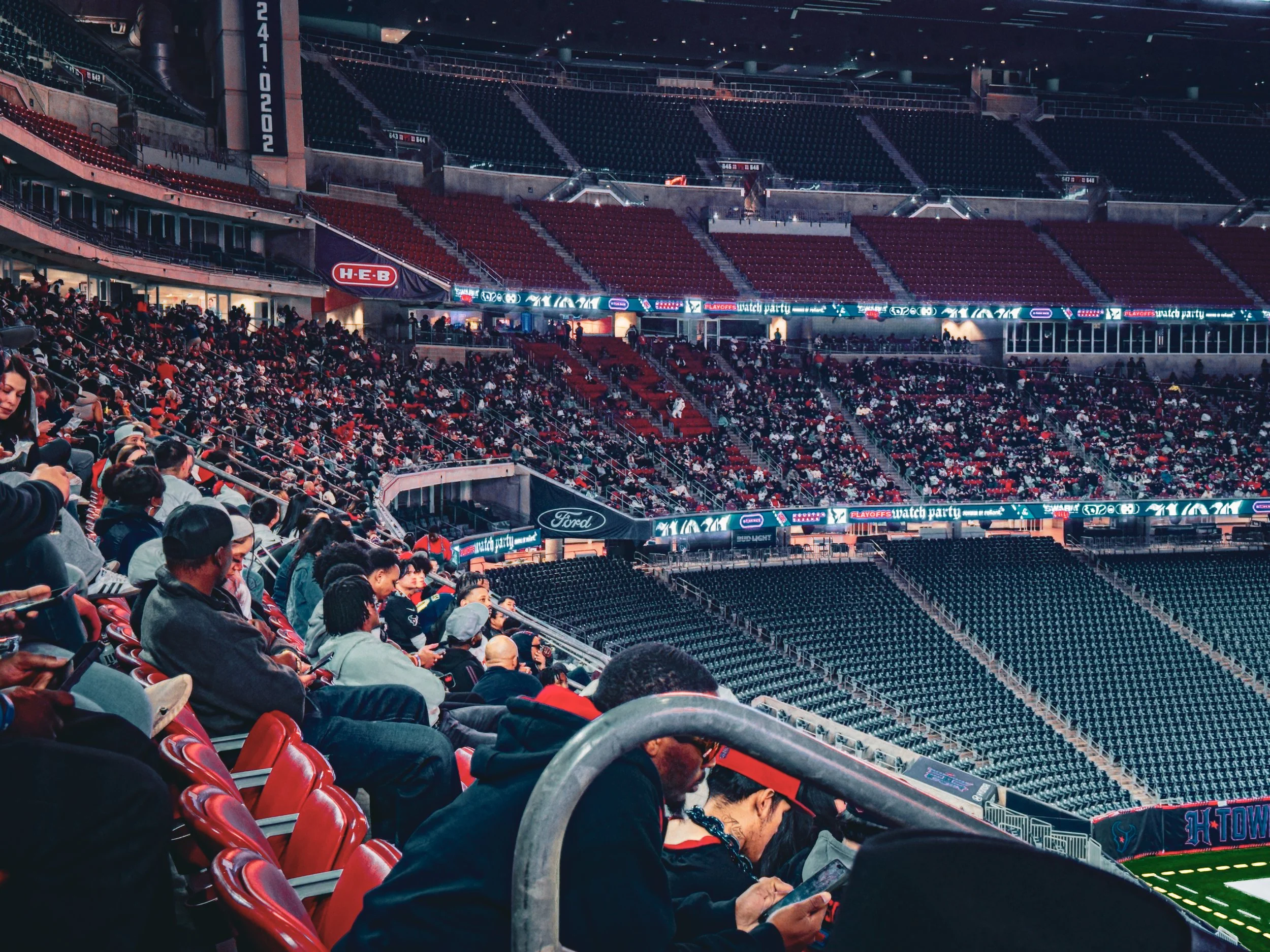 People sitting in stadium seats before a sporting event or concert, with numerous empty seats and electronic banners displaying advertisements.