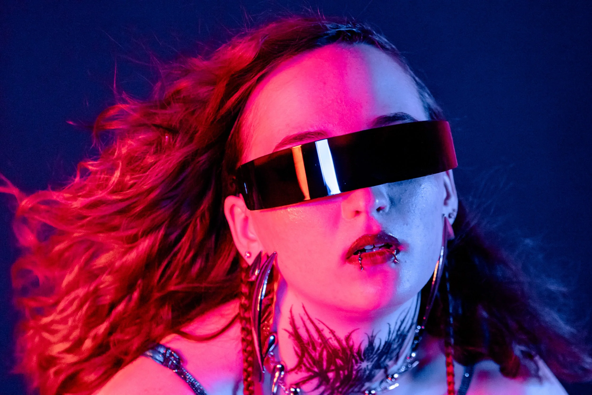 A young woman with wavy hair wearing large, black visor sunglasses, with piercings, tattoos, and jewelry, illuminated by vibrant pink and blue lighting.