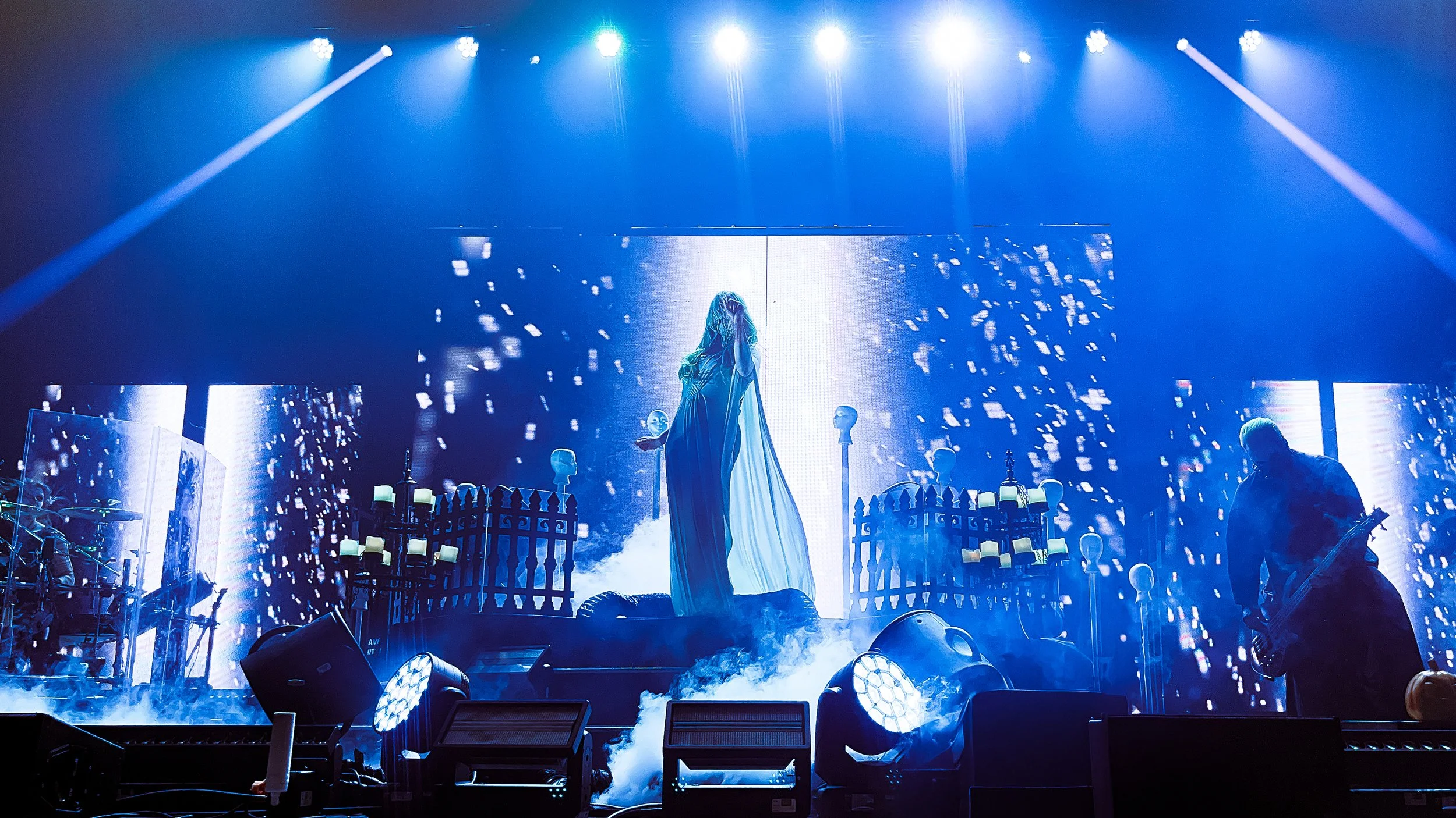 Stage with a female singer in a flowing dress, surrounded by blue lighting, fog, and LED screens displaying abstract visuals. Musicians and equipment are visible around the stage.