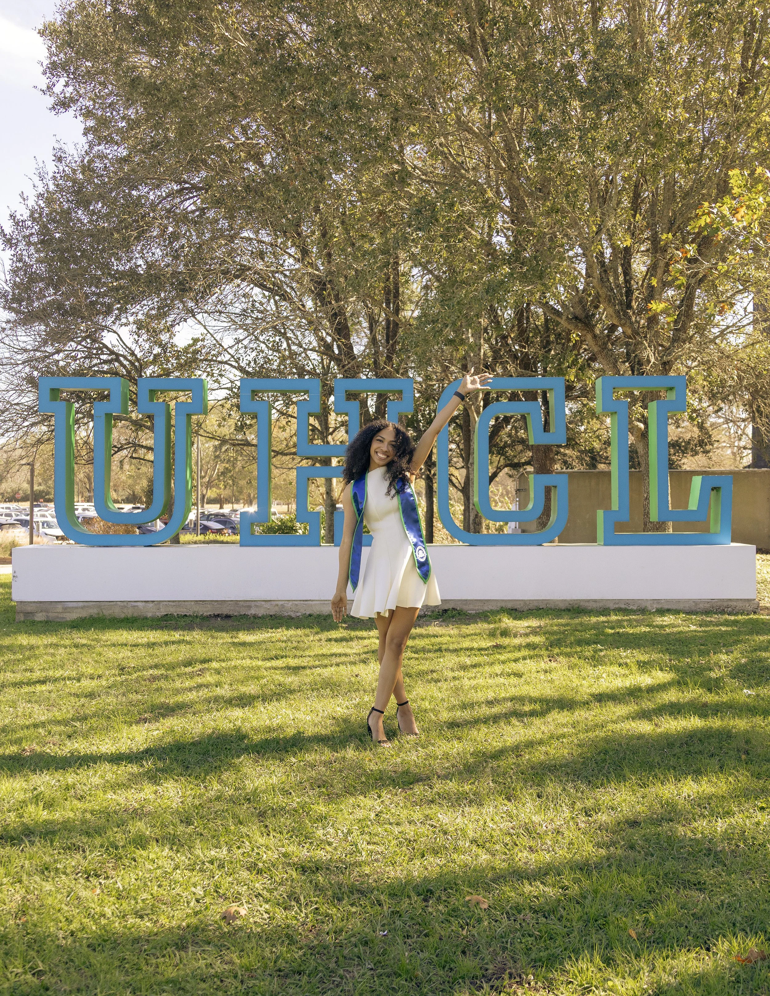 A young woman in a white dress and high heels standing in front of a large university sign that reads 'UICL'. She is smiling and pointing upward with her right hand, with a graduation stole around her neck, and trees and parked cars are visible in th