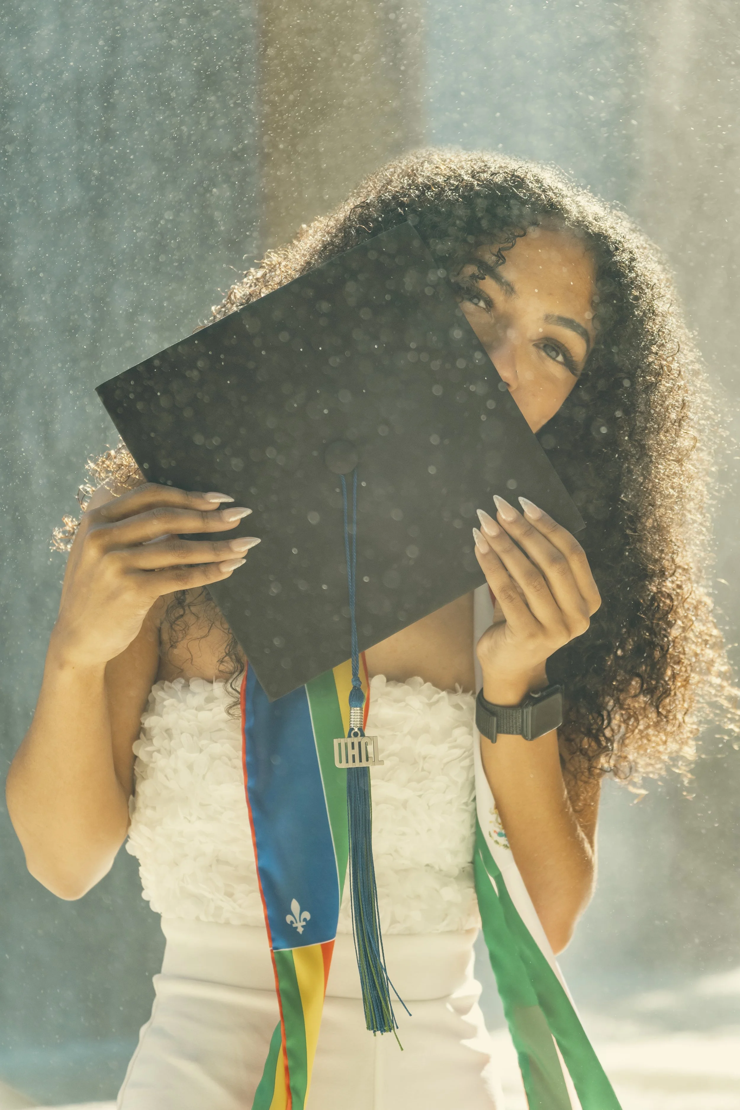 Young woman holding her graduation cap in front of her face outdoors during daytime, with sunlight shining through her curly hair, wearing a white dress and a black watch.