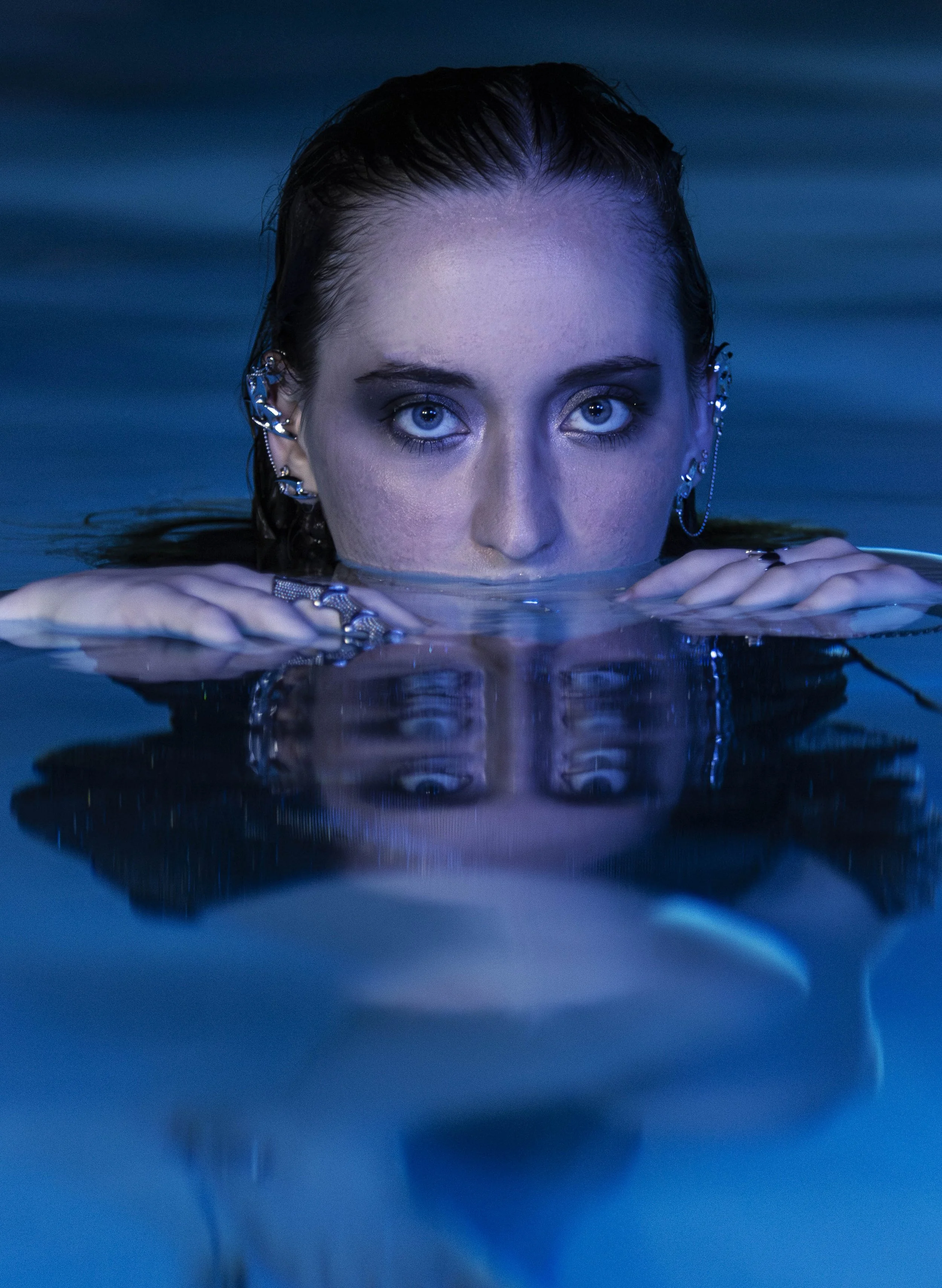 A woman with bright blue eyes and earrings is partially submerged in water, with only her face and hands visible above the surface, reflecting on the water.
