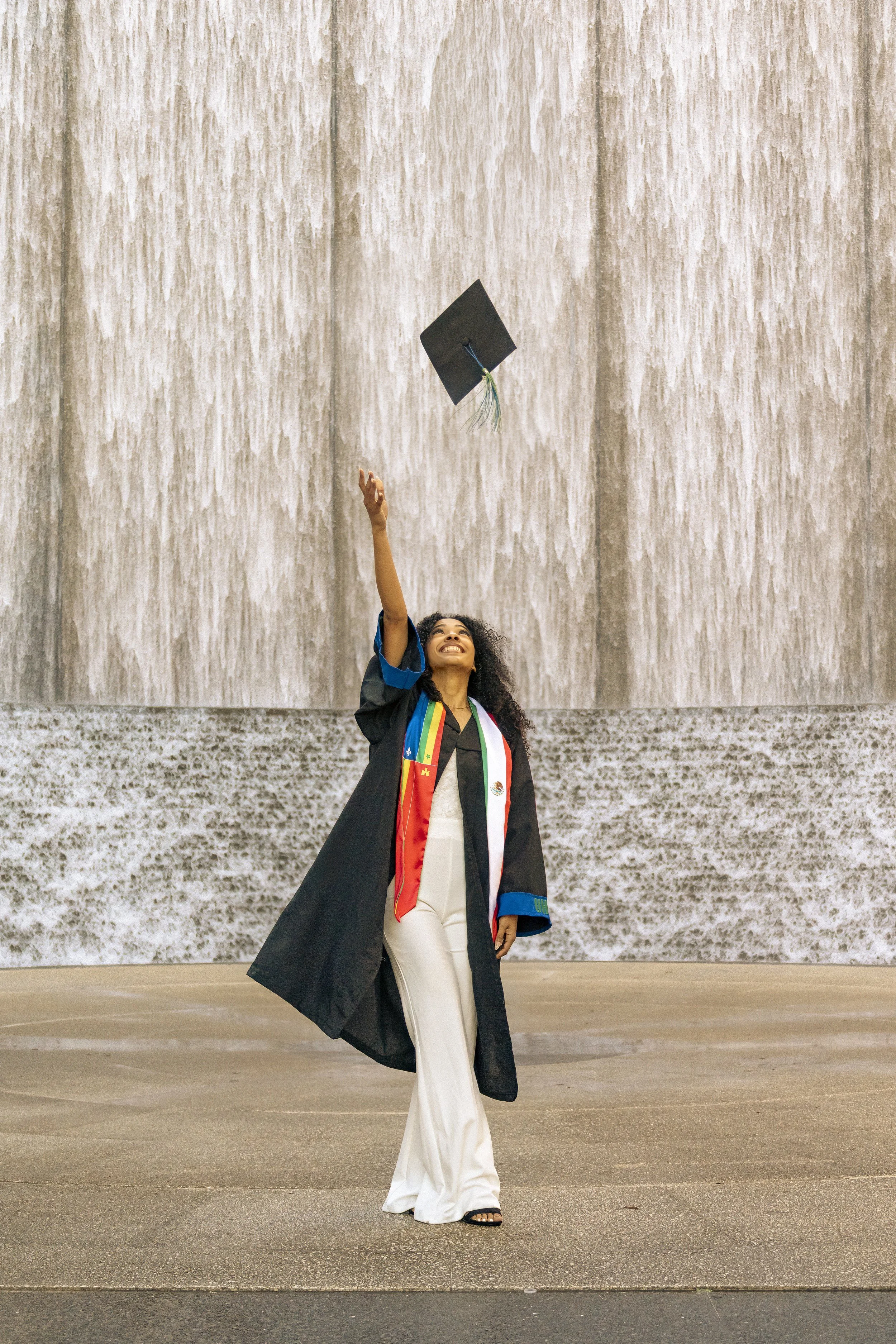 A woman in graduation attire throws a mortarboard cap into the air in front of a large stone wall.