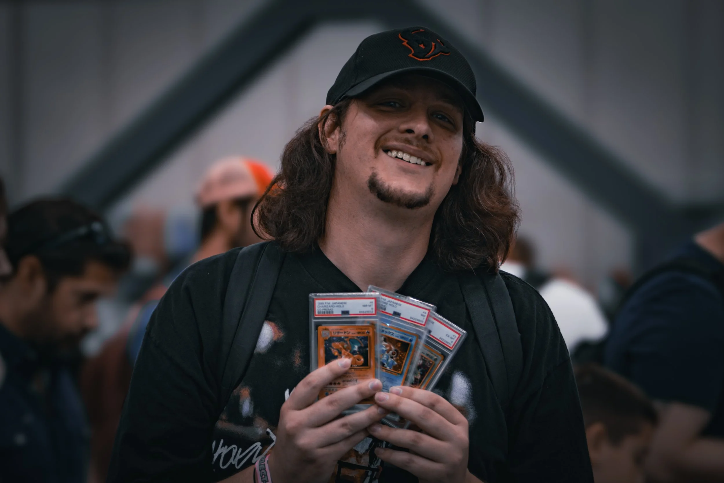 A man with long brown hair and a goatee smiling and holding several Pokémon cards in a busy, indoor Pokémon event.