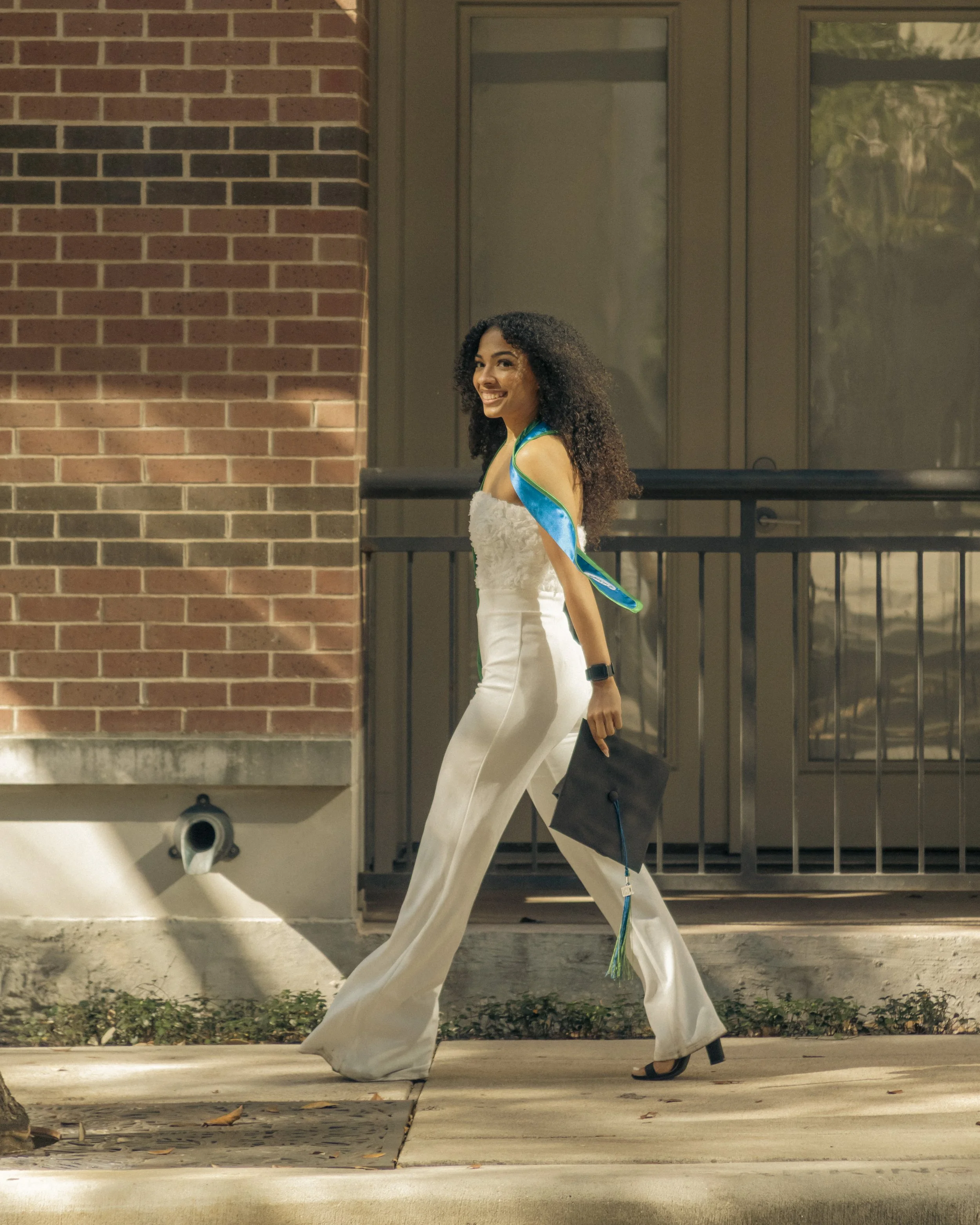 A young woman in a white dress and black heels is walking on a sidewalk, holding a graduation cap, with a big smile.