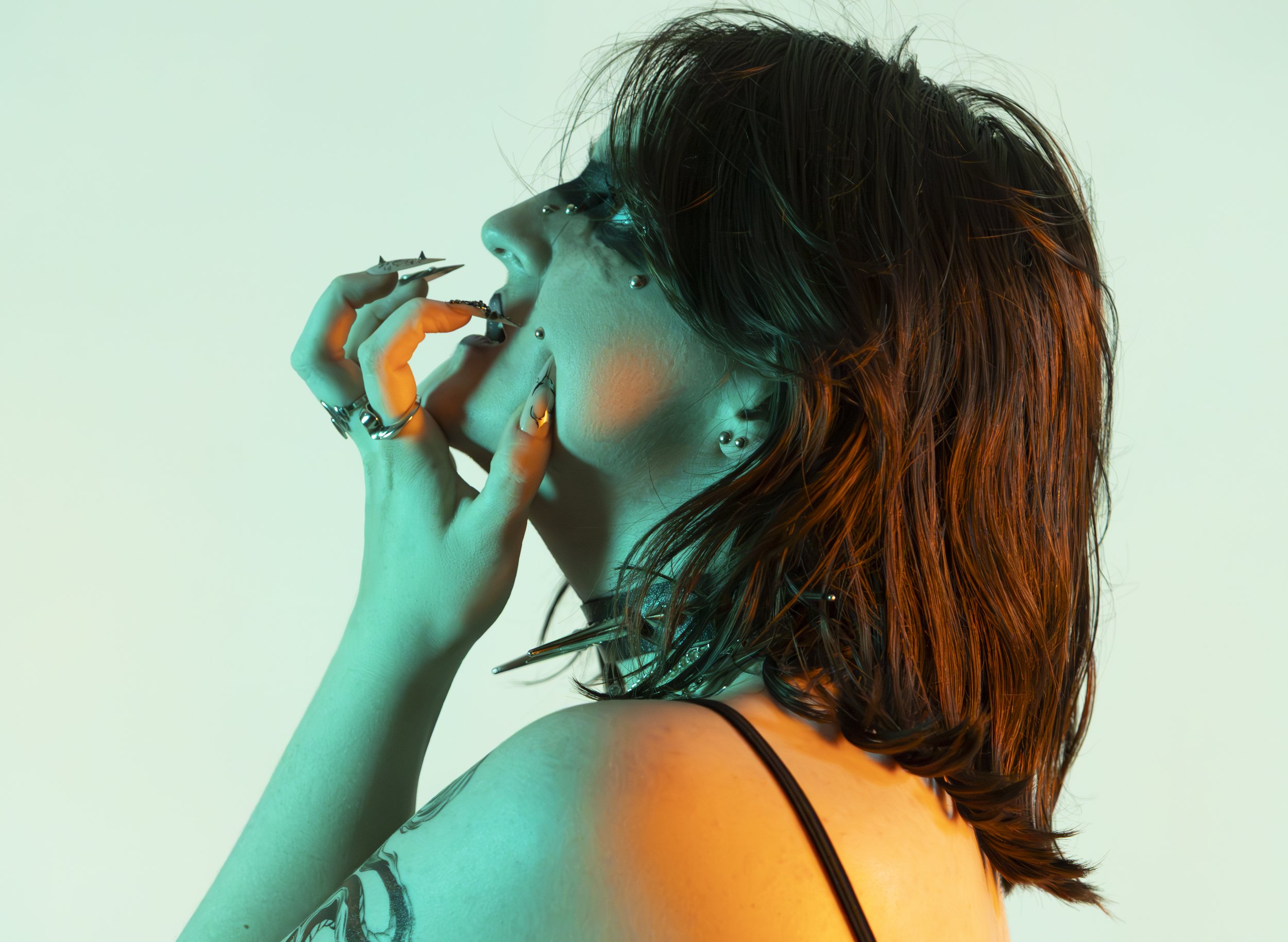 A woman with shoulder-length, reddish-brown hair, multiple facial piercings, and tattoos, holding a cigarette near her mouth, with colorful lighting in the background.