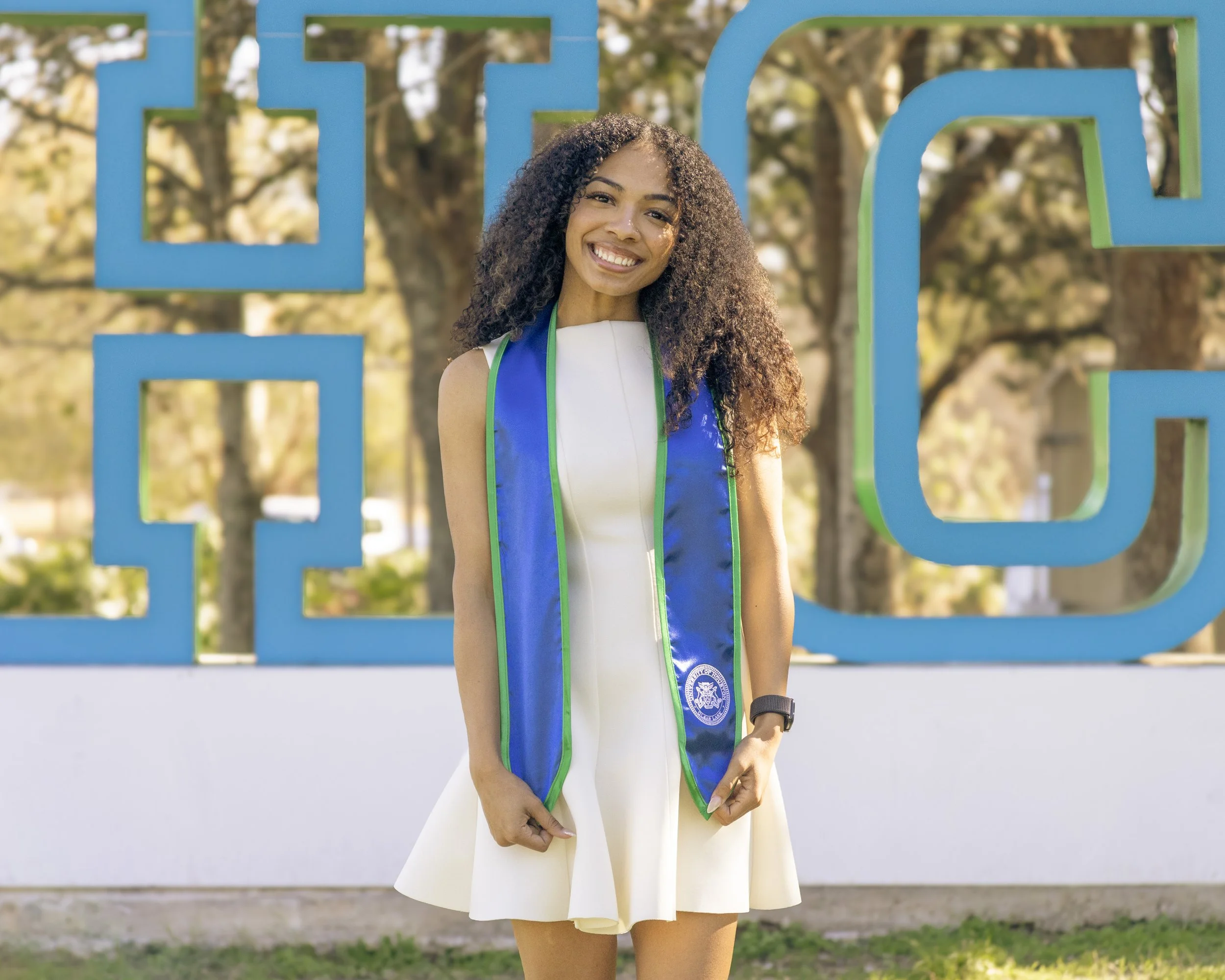 A young woman in a white sleeveless dress and a blue graduation sash with green trim, smiling and standing outdoors in front of large blue letters on a white wall, with trees in the background.