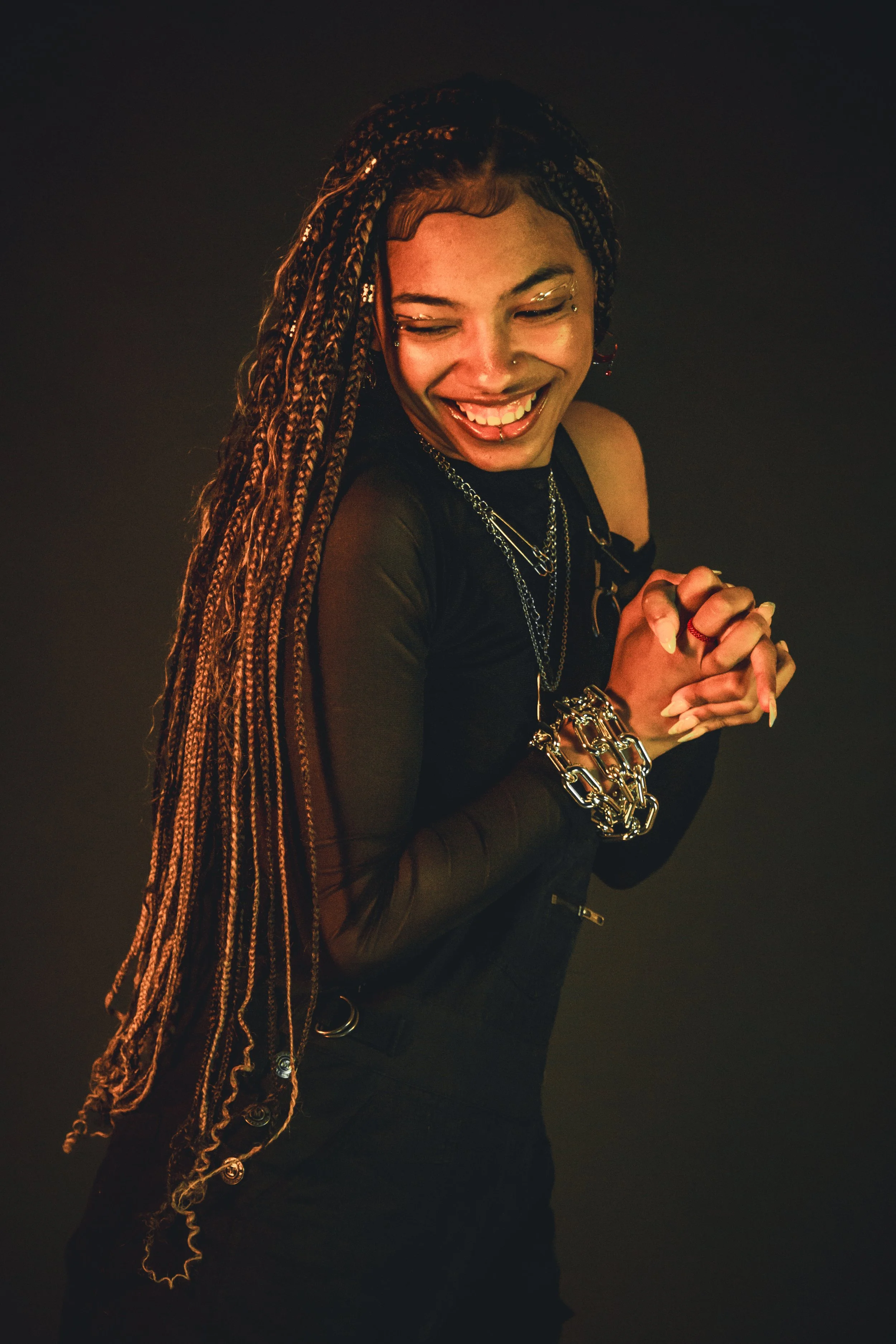 A woman with braided hair, smiling and clasping her hands together, wearing a black top with sheer sleeves, layered necklaces, and chunky jewelry, against a dark background.