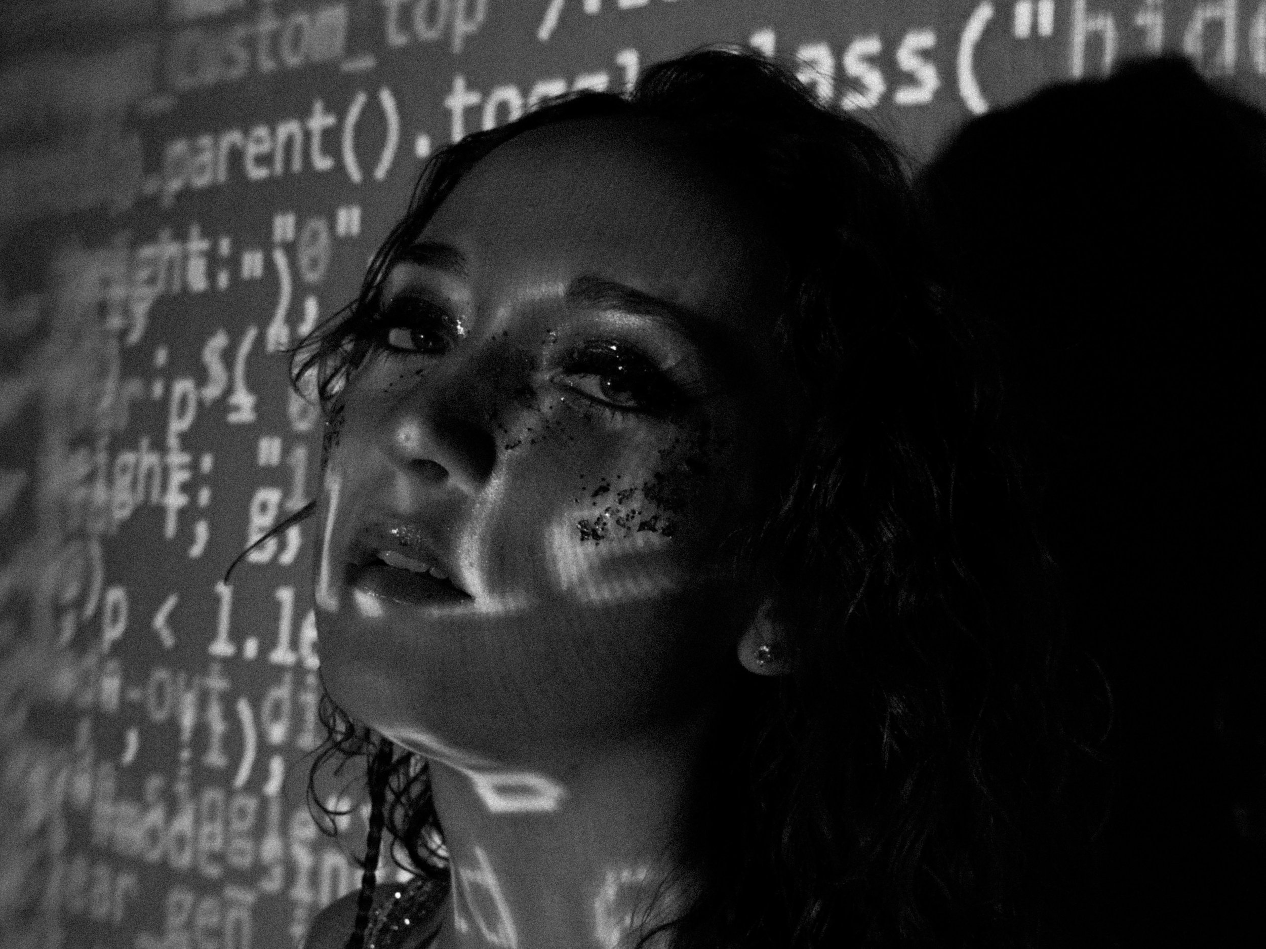 A woman with curly hair and makeup looking at a projection of code on her face and background.