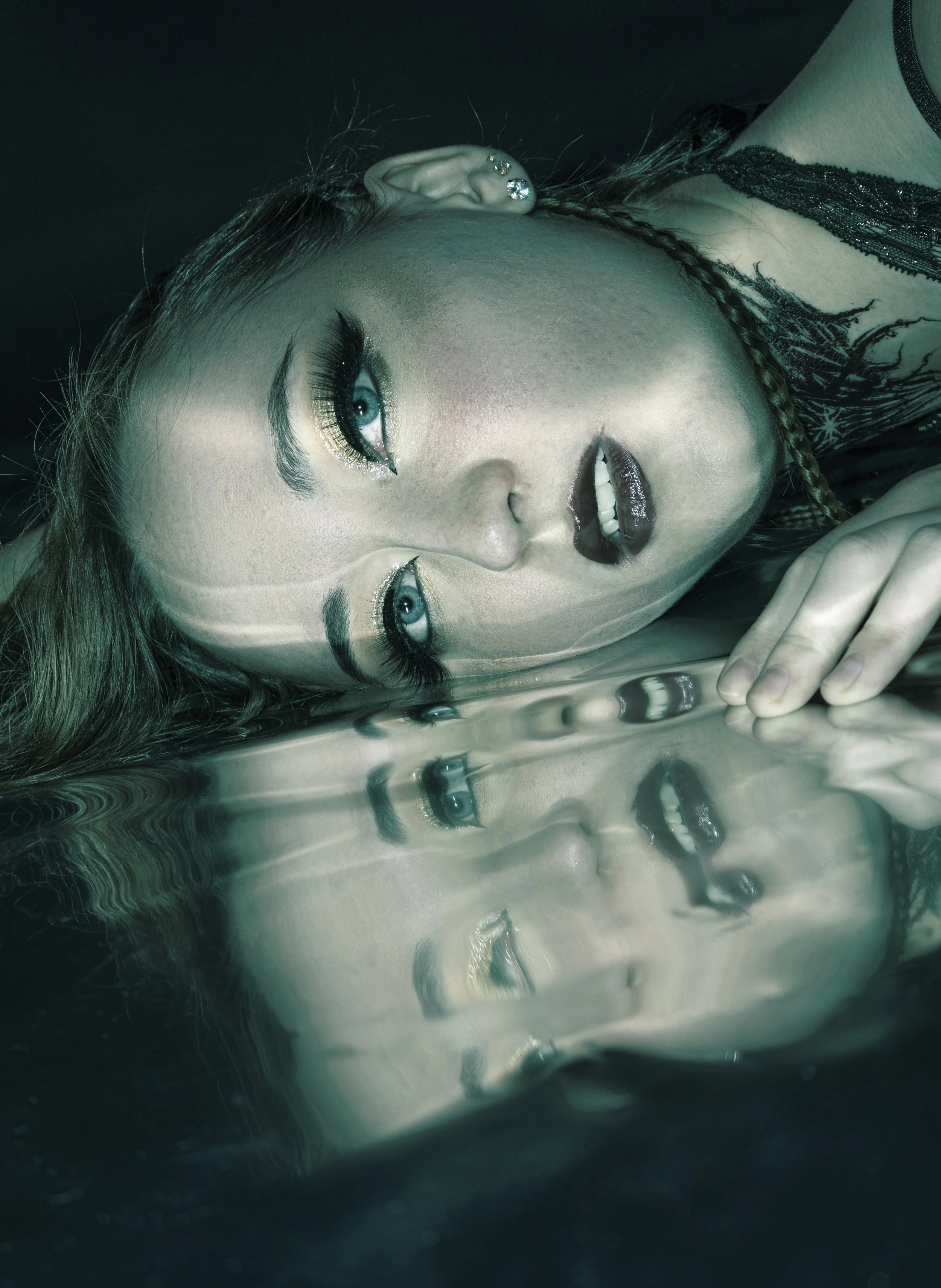 A woman with dramatic makeup and jewelry lying down with her face close to a reflective surface, creating a mirror image of her face.