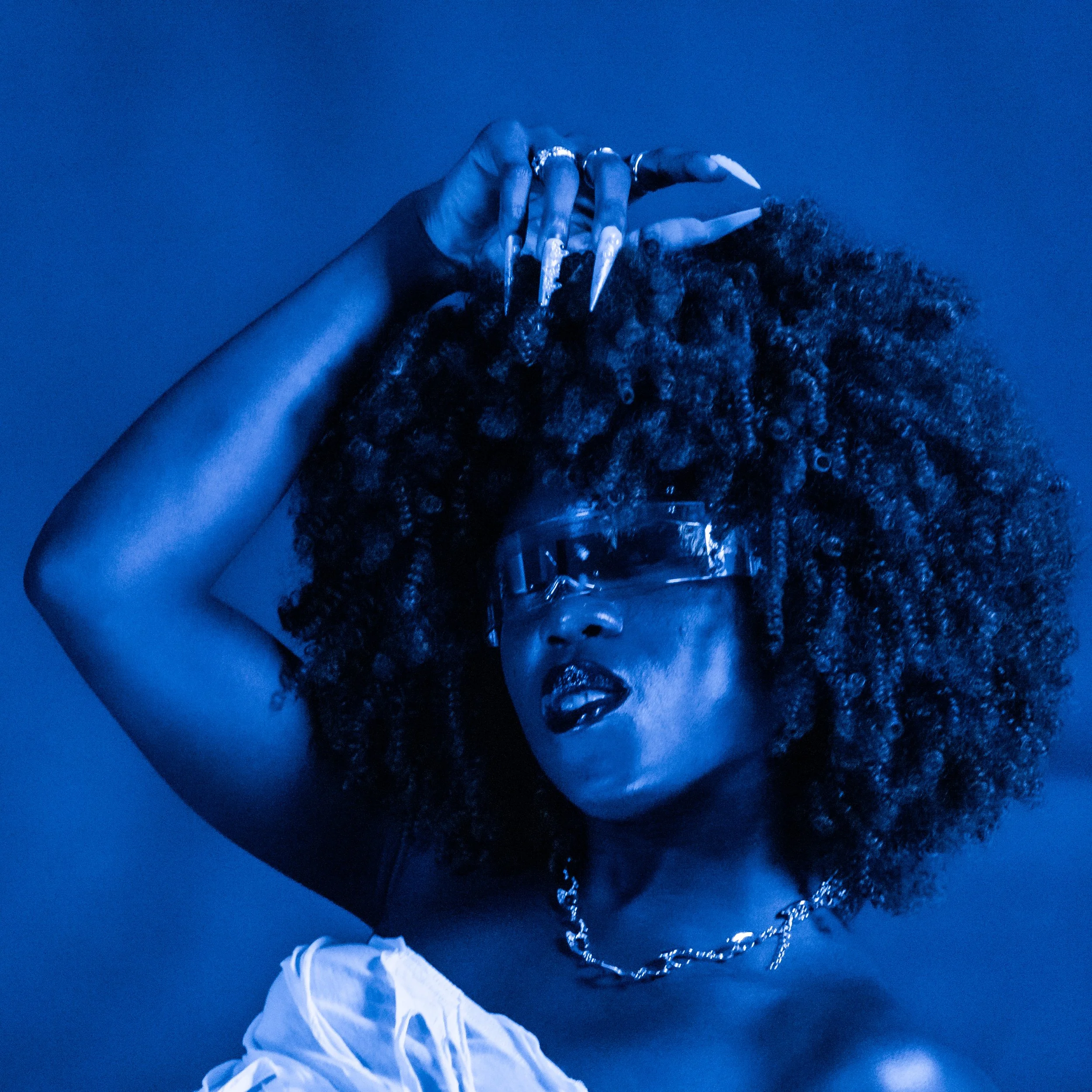 A woman with voluminous curly hair, wearing fashion sunglasses, a white off-shoulder top, chunky chain necklace, and shiny lipstick, posing against a blue background with her hand above her head.
