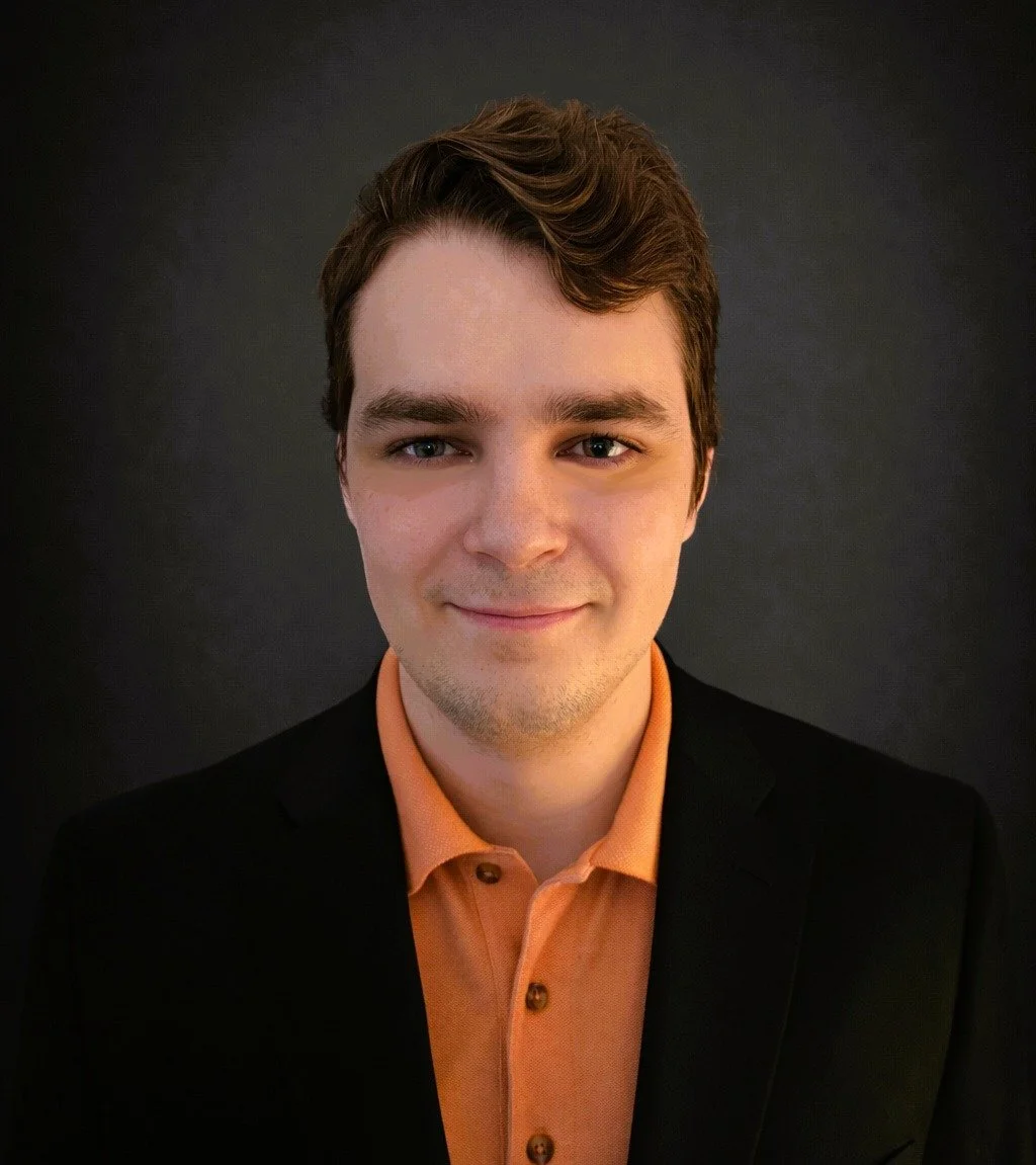 Portrait of Adam Galauner with brown hair, wearing an orange shirt and black jacket, smiling against a dark background.