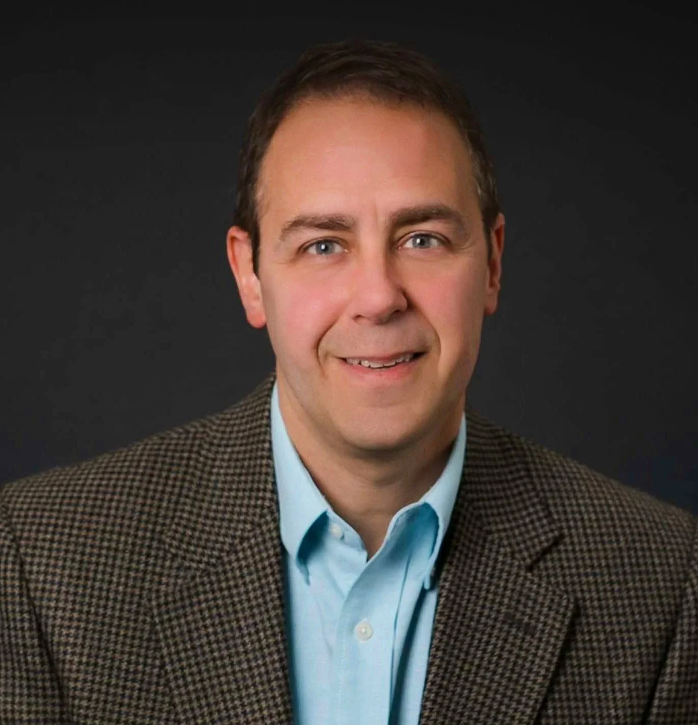 Headshot of Mark Galauner with short brown hair, wearing a light blue collared shirt and a brown checkered blazer, against a dark background.
