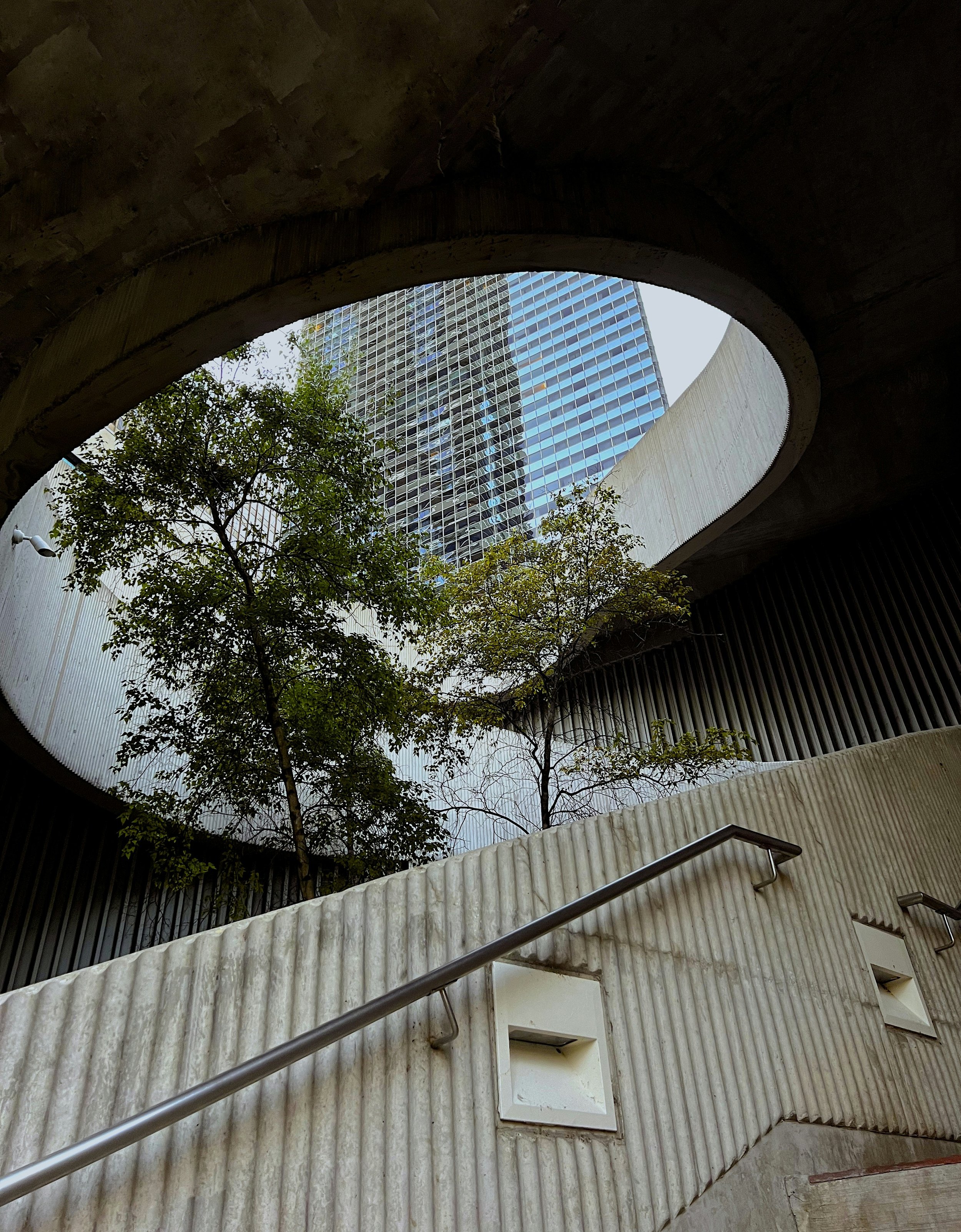 View from below of a modern high-rise glass building in Chicago seen through a circular opening in a concrete structure with trees below and a staircase railing in the foreground.