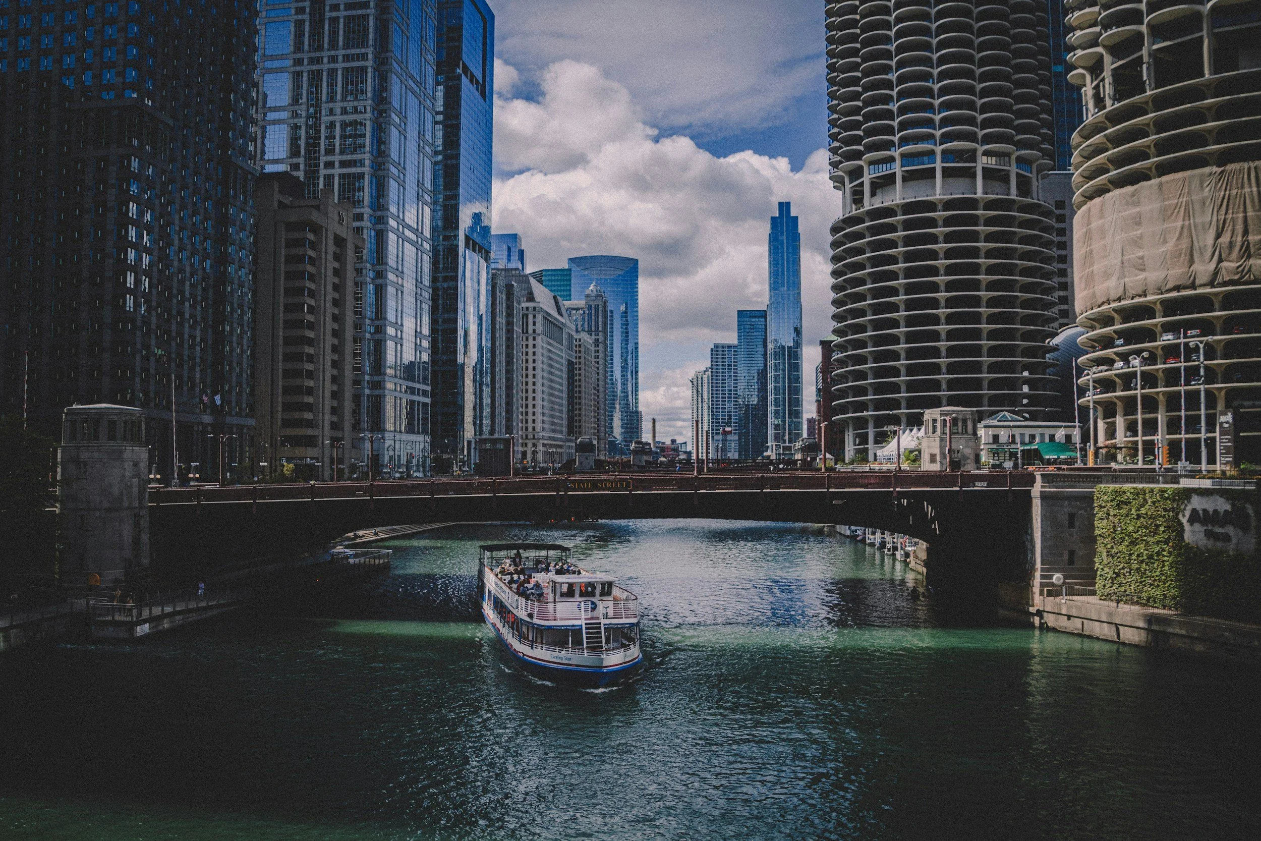 A dramatic photo of Chicago featuring tall modern glass buildings and unique architectural high-rises, with the Chicago River running through the downtown area and a boat cruising on the water. The sky is partly cloudy.