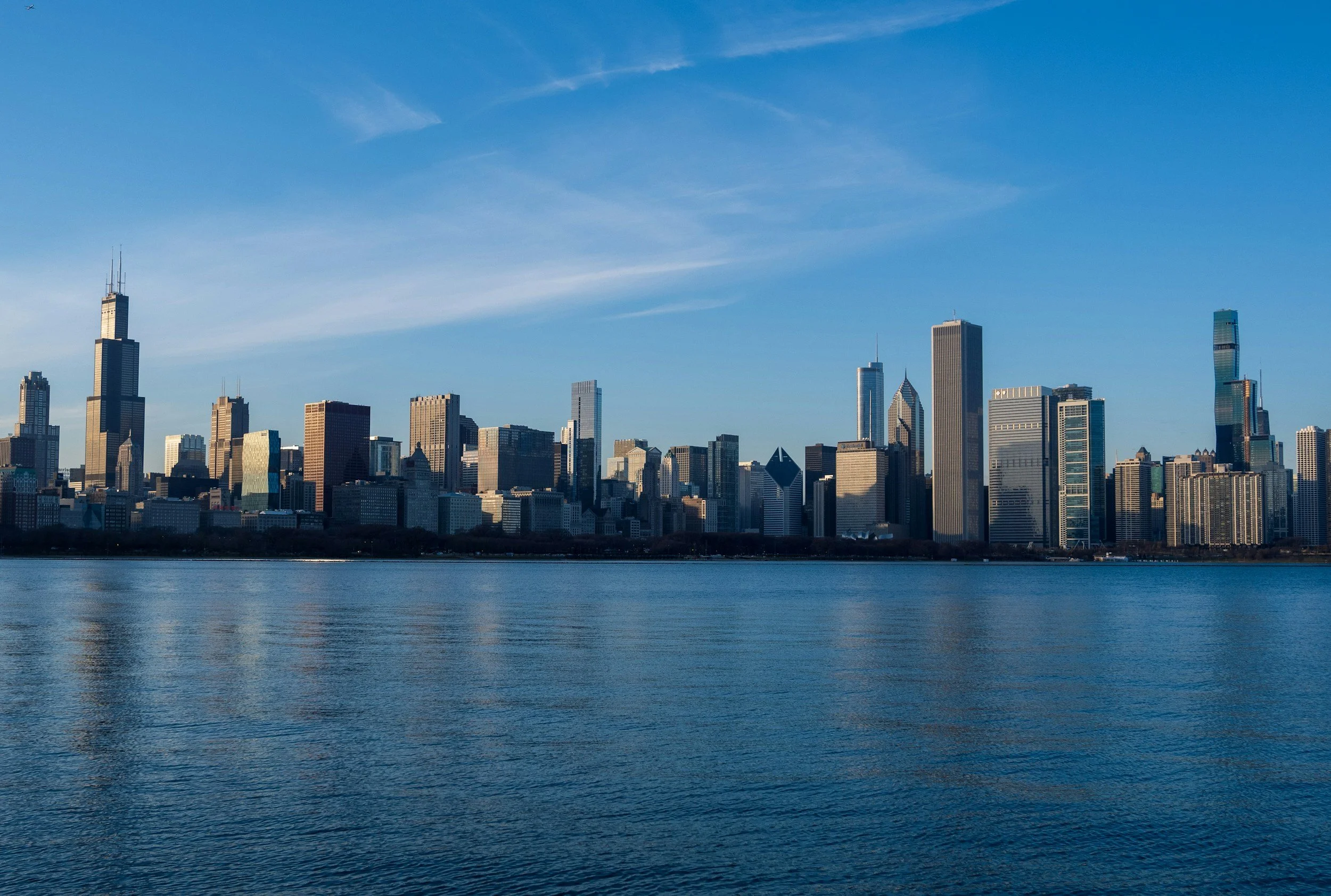 The Chicago skyline with tall skyscrapers across a body of water under a blue sky with some clouds.