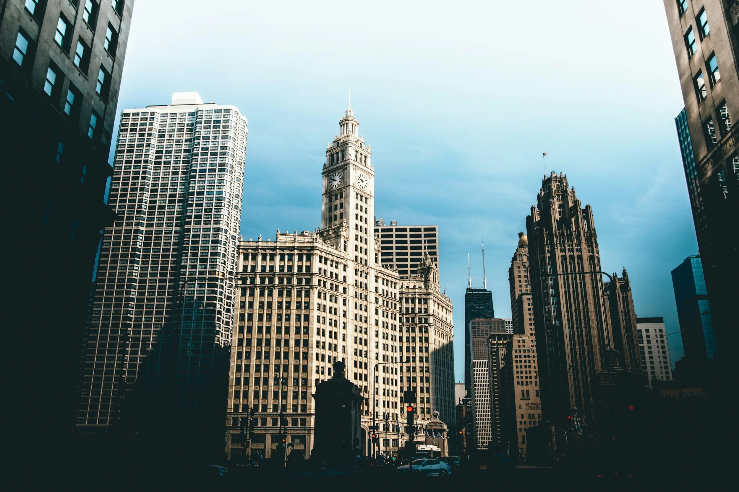A Chicago cityscape featuring tall skyscrapers, including a clock tower with a white façade and black clock faces, against a blue sky with some clouds.
