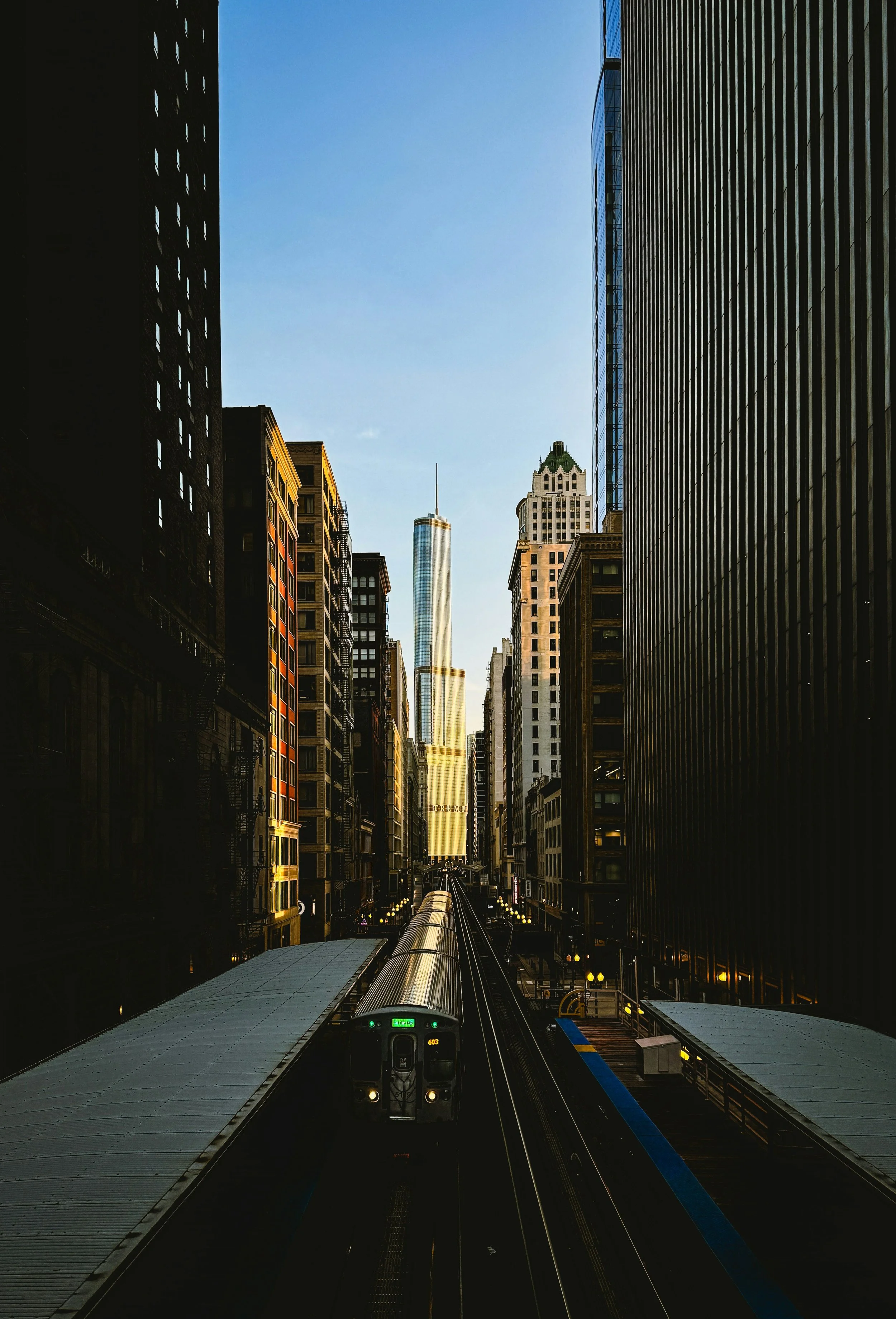 A Chicago city street view with train tracks running through the middle, tall buildings on both sides, and a modern skyscraper in the background under a clear blue sky.