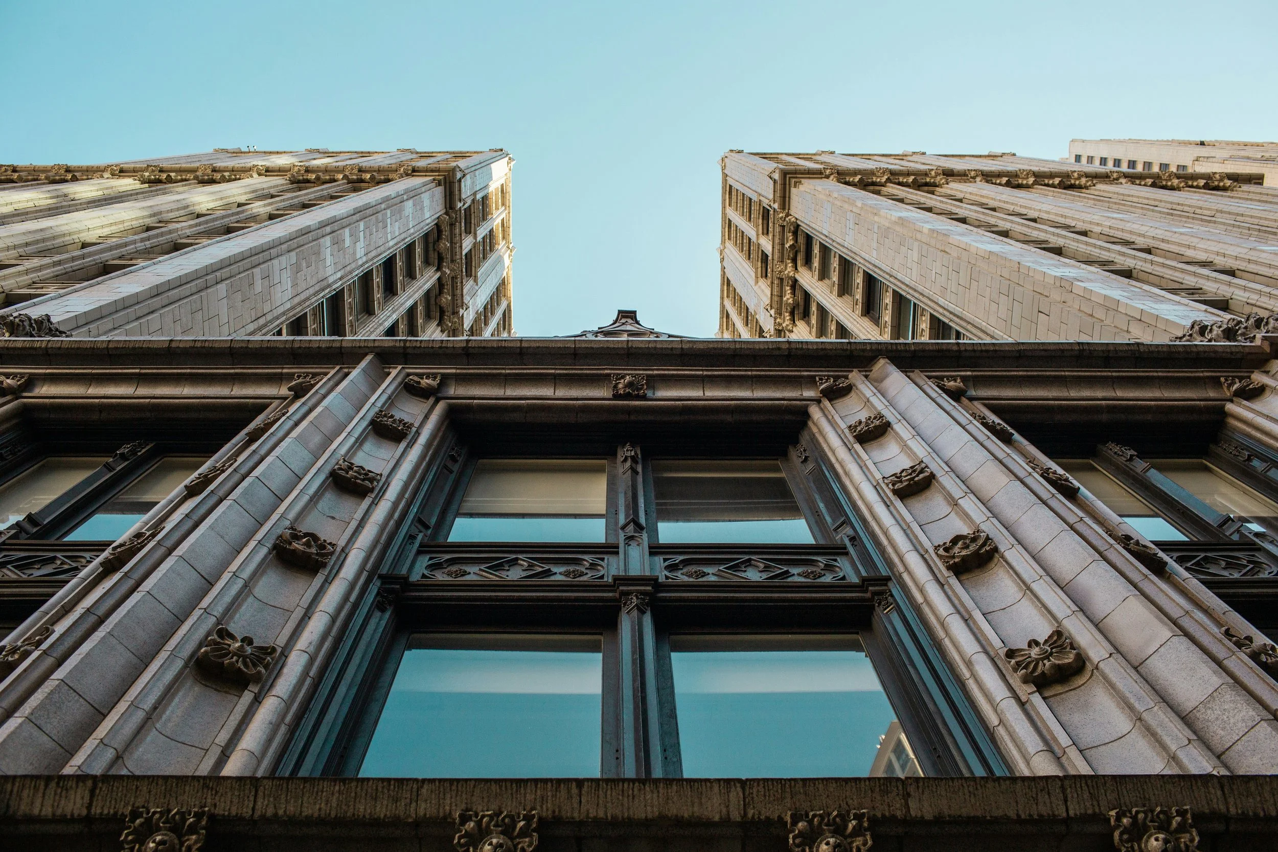 Looking up at the facades of two tall, ornate skyscrapers in Chicago, with detailed stonework and large windows, converging towards the clear blue sky.