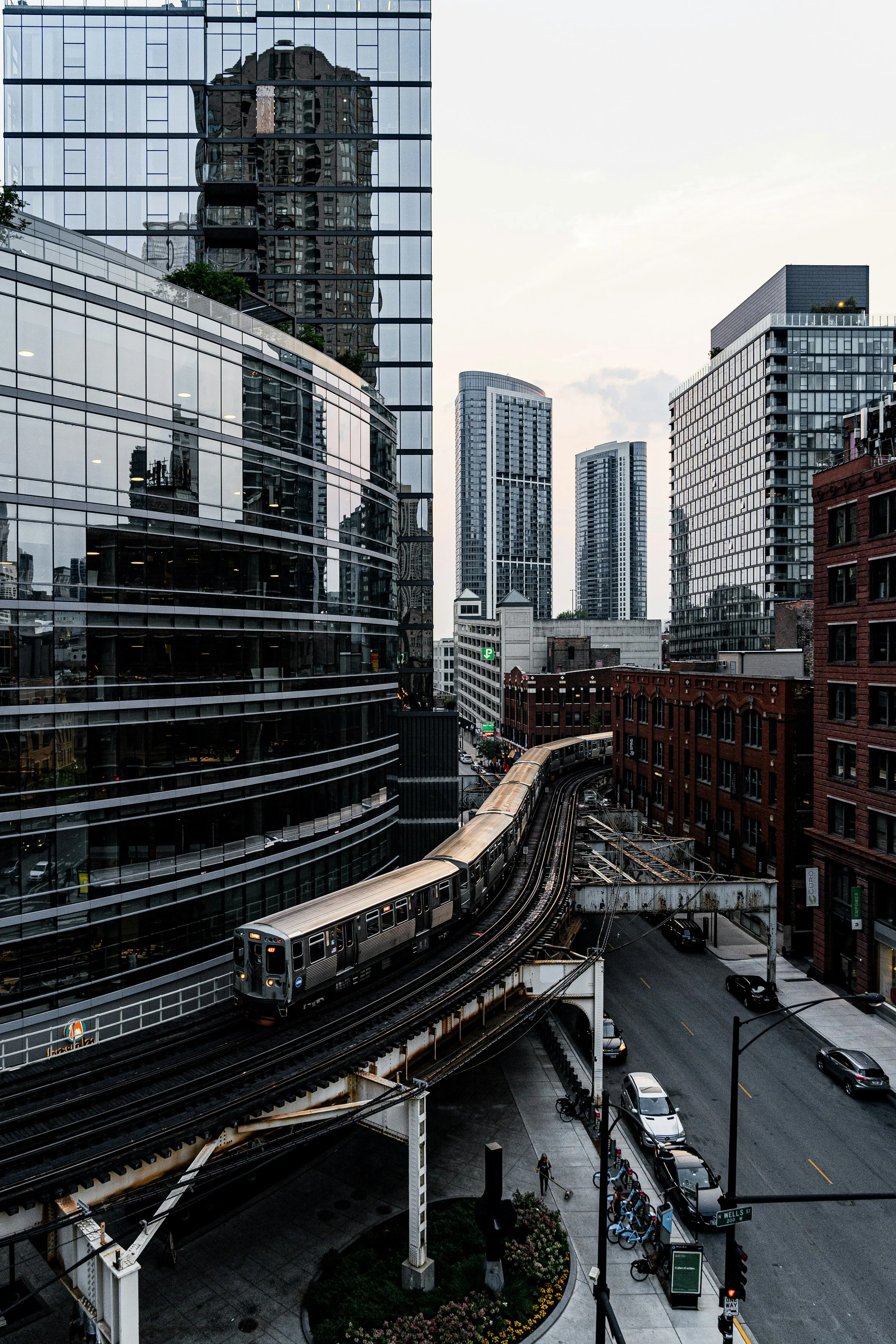 Urban cityscape in Chicago with high-rise glass buildings, trains on elevated tracks, and a street with cars and pedestrians at dusk.