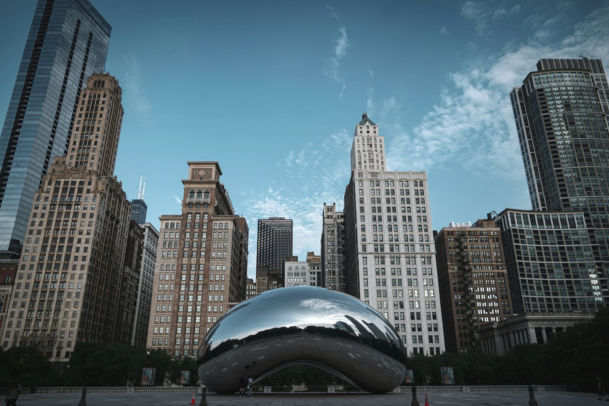 Two people standing in front of the reflective sculpture at Millennium Park, with tall skyscrapers in the background under a partly cloudy sky.
