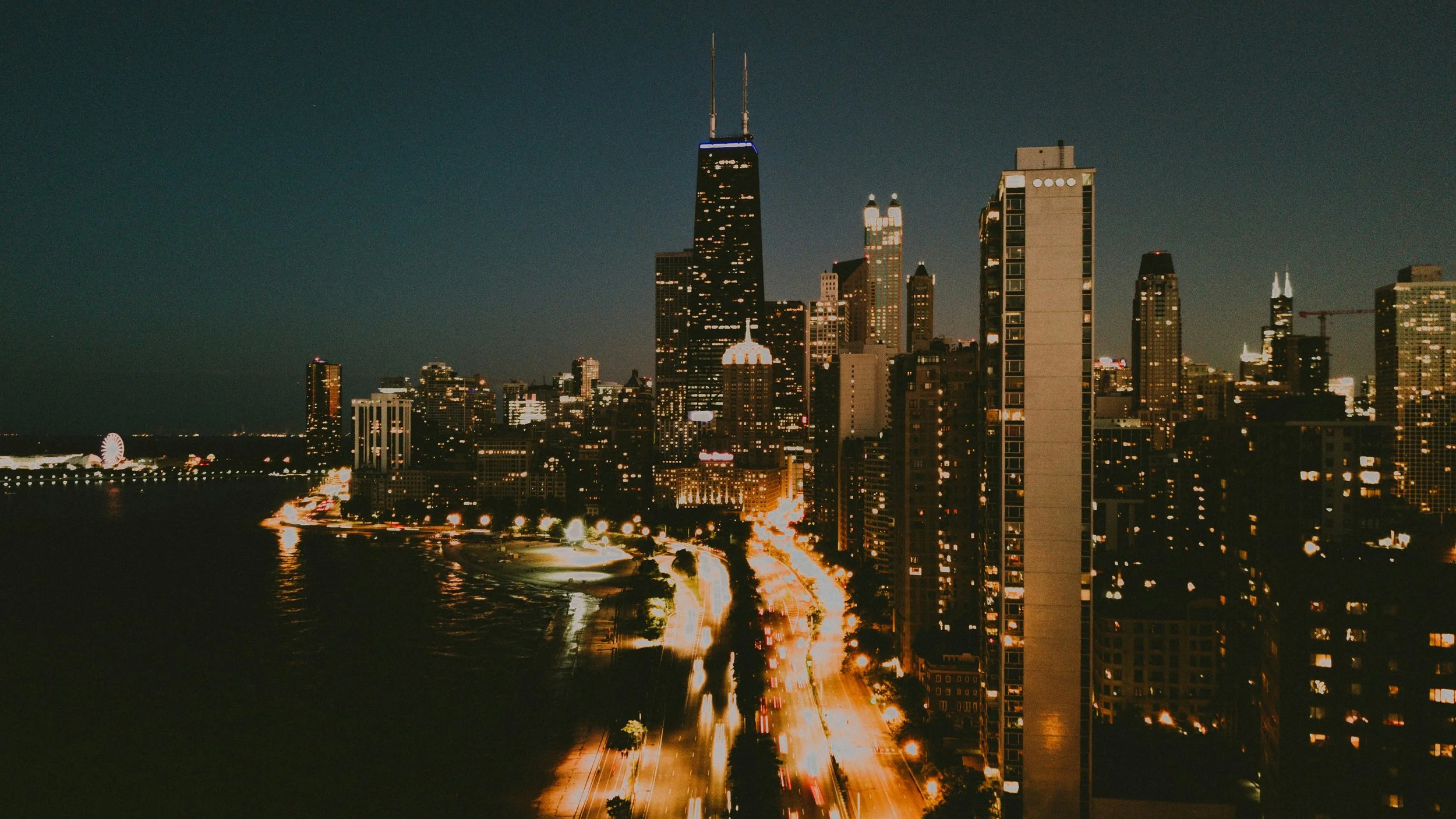 Nighttime cityscape of the Chicago, Illinois, skyline featuring tall skyscrapers, including a building with twin antennas, with illuminated streets and reflections on Lake Michigan.