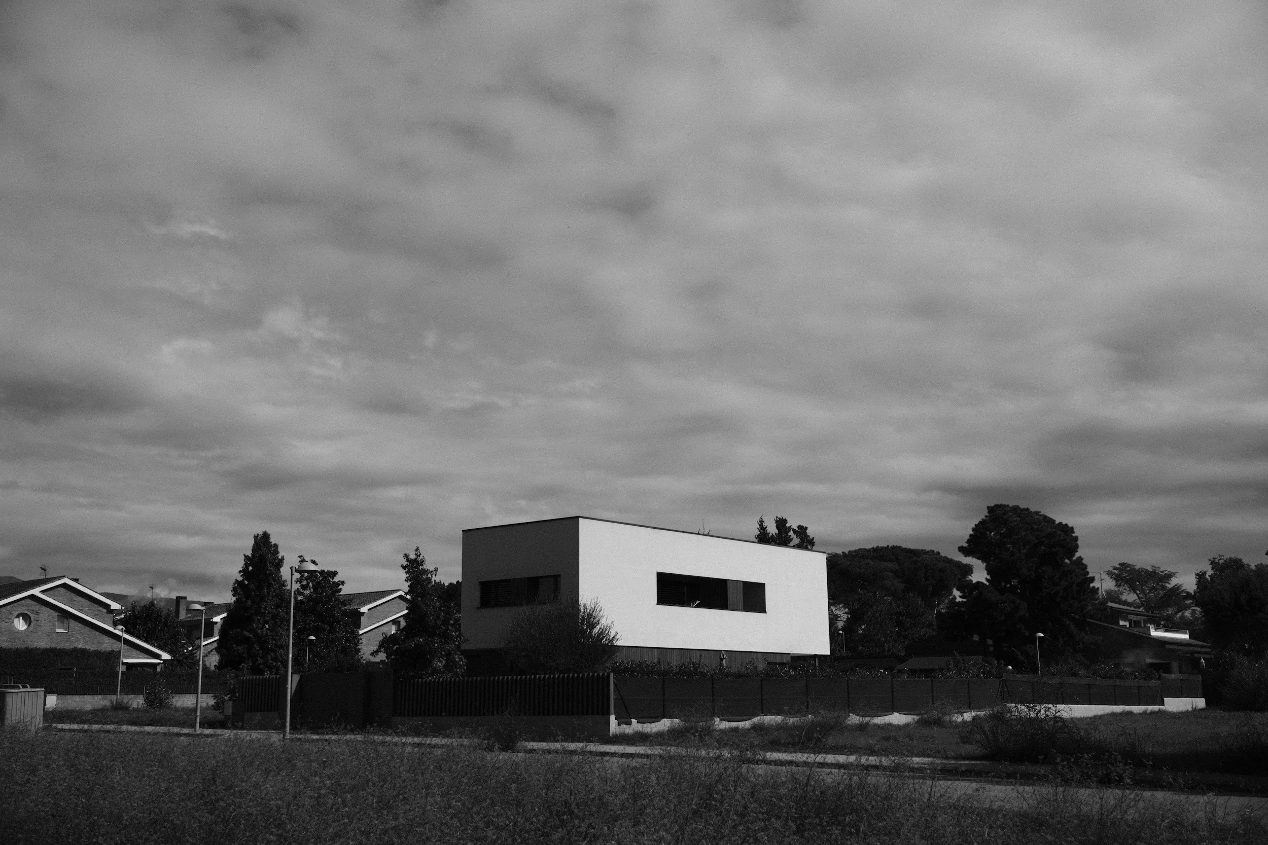 Modern white rectangular house with large horizontal window, surrounded by trees in a suburban area under a cloudy sky, in black and white.