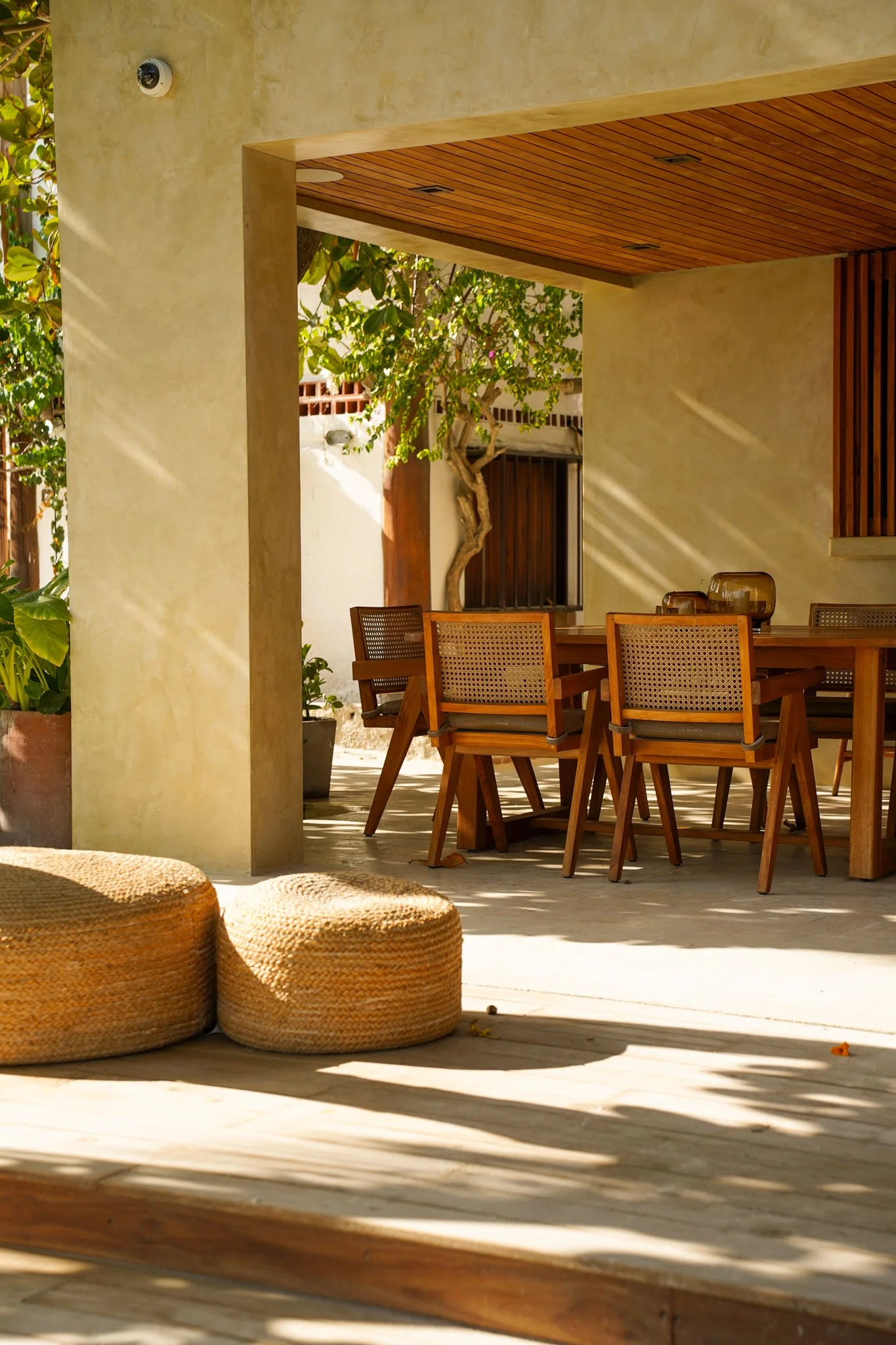 An outdoor patio area with a wooden dining table and chairs, potted plants, and woven ottomans, with sunlight casting shadows.