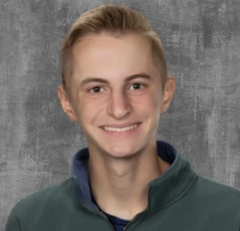 A young man with blonde hair smiling, wearing a green jacket over a dark shirt, standing against a gray background.