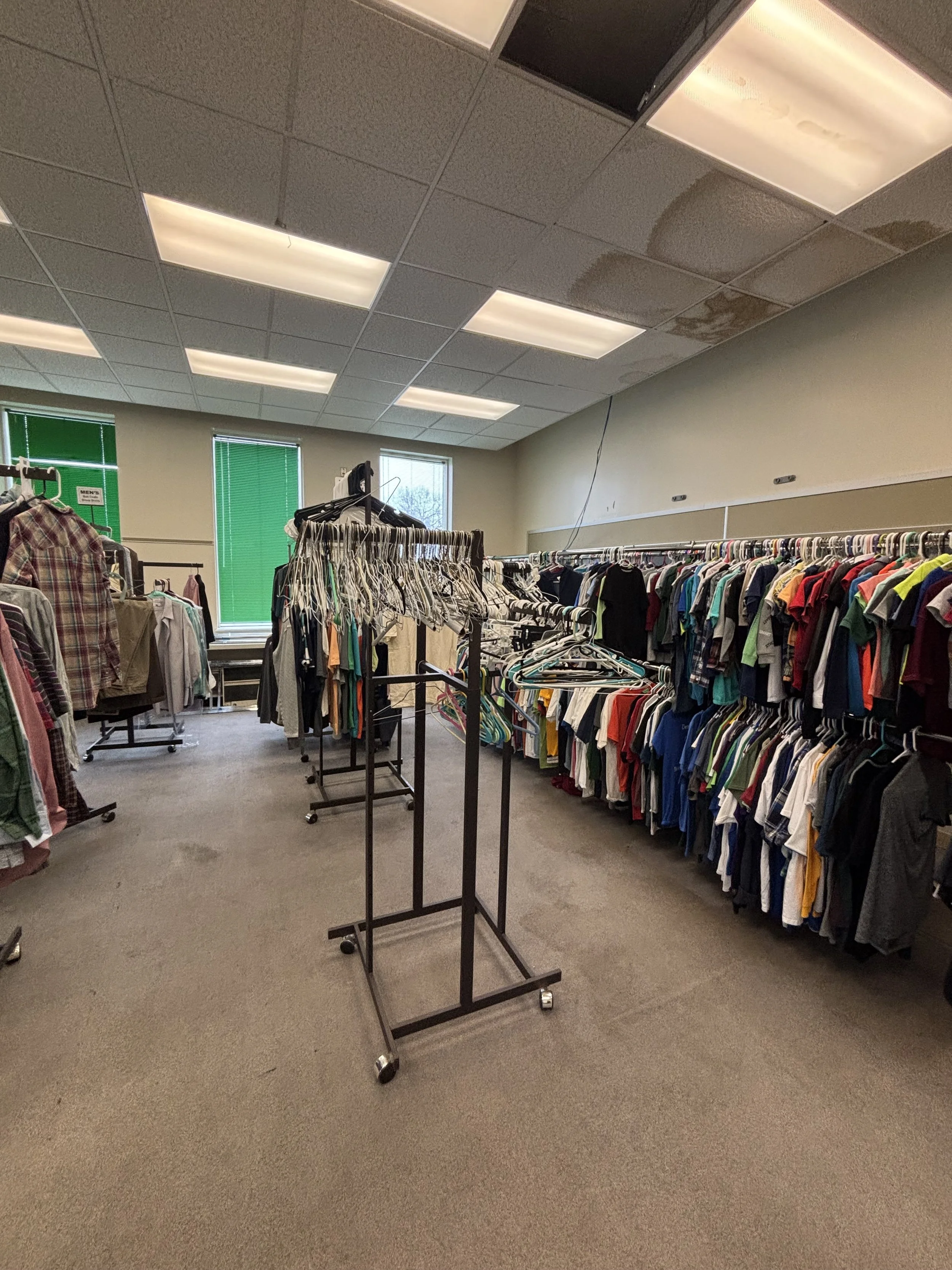 A clothing store with racks of clothes, windows with green blinds, and a beige carpeted floor. There are fluorescent ceiling lights and a black portable clothing rack on wheels in the center.