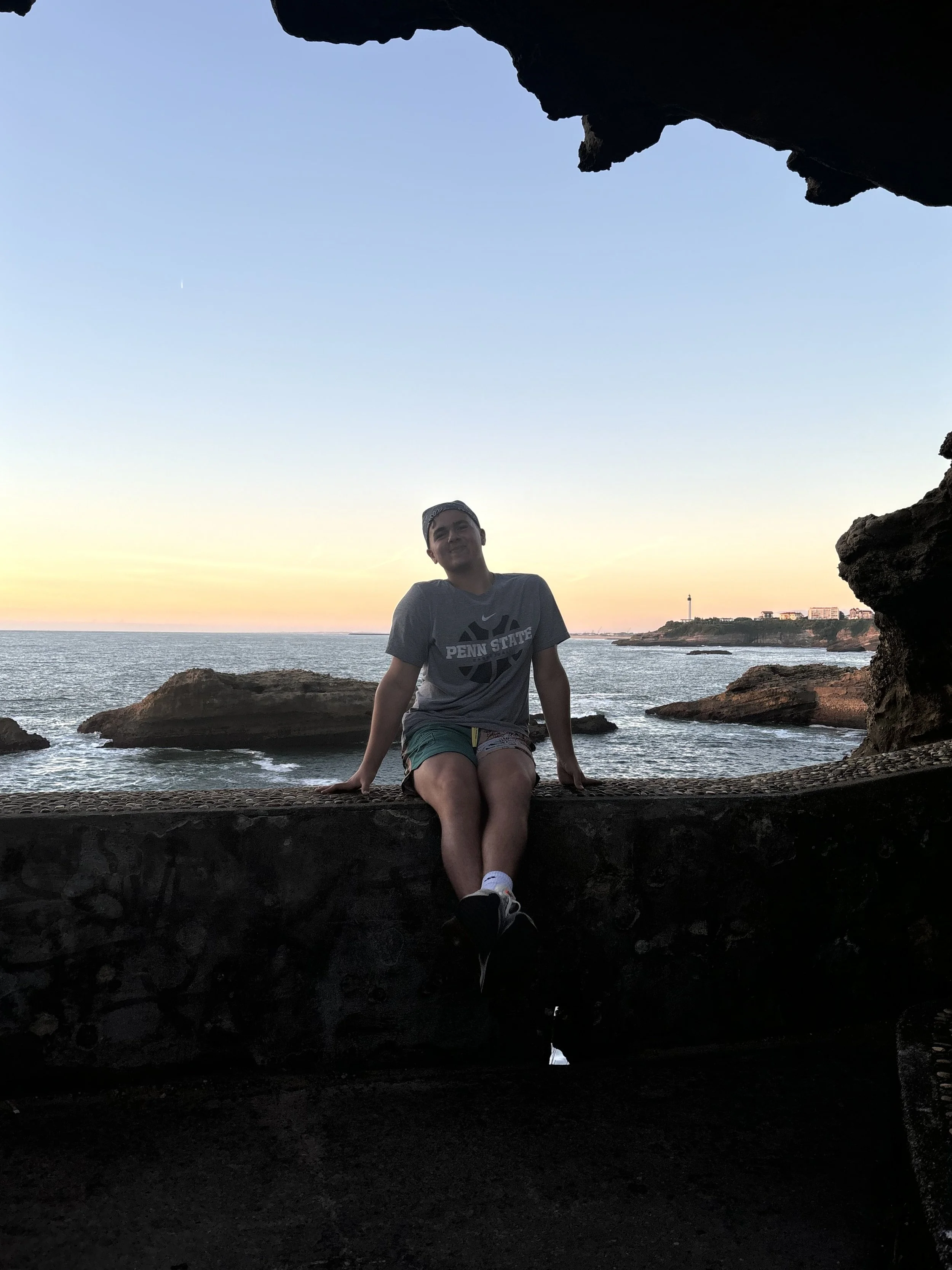 A man sitting on a ledge with a sunset over the ocean behind him, viewed from inside a dark cave or tunnel with an opening at the top.