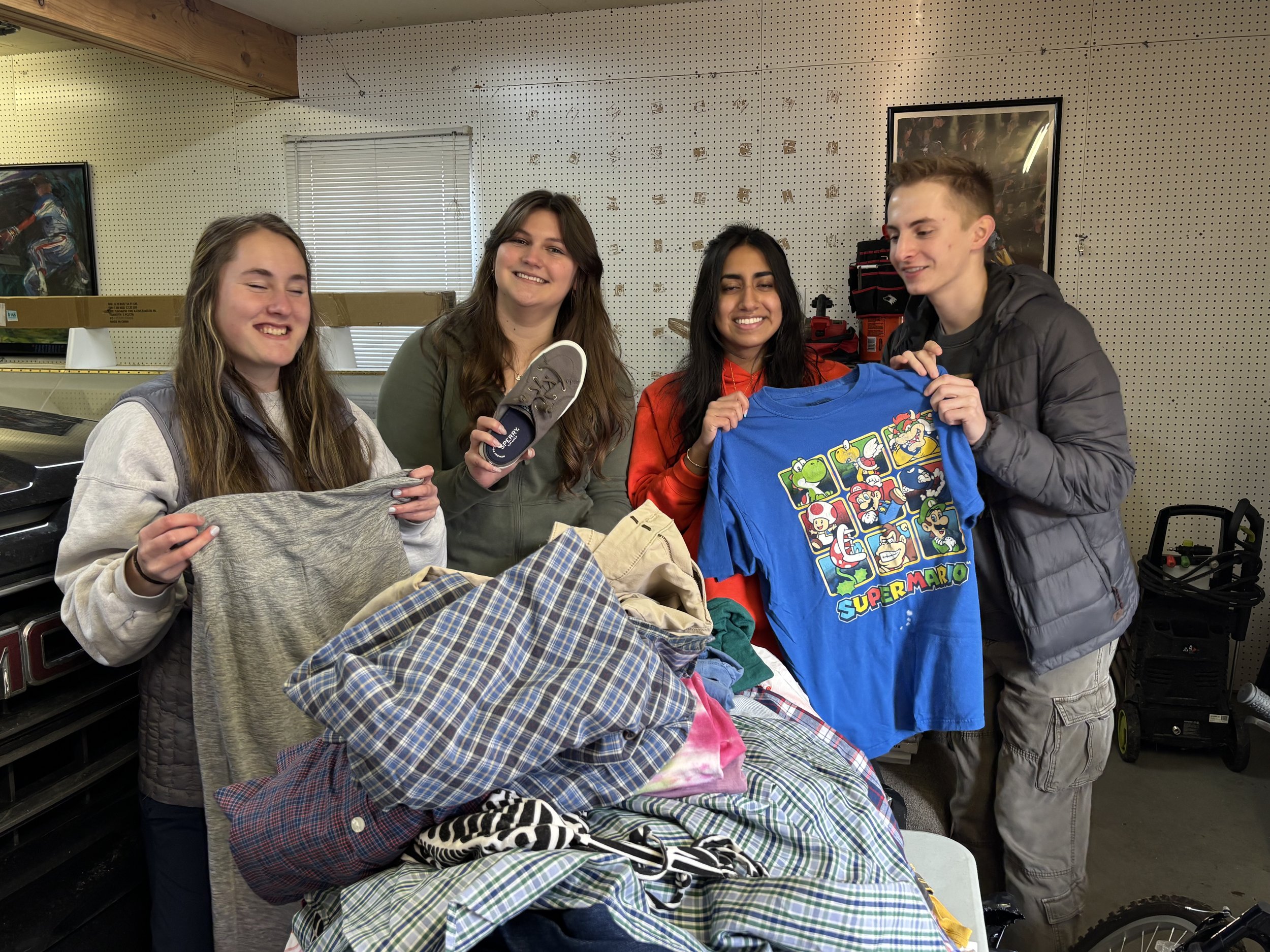 Four smiling young people standing behind a table with colorful clothes, holding a blue Super Mario shirt and a gray shoe, in a room with pegboard walls and framed posters.