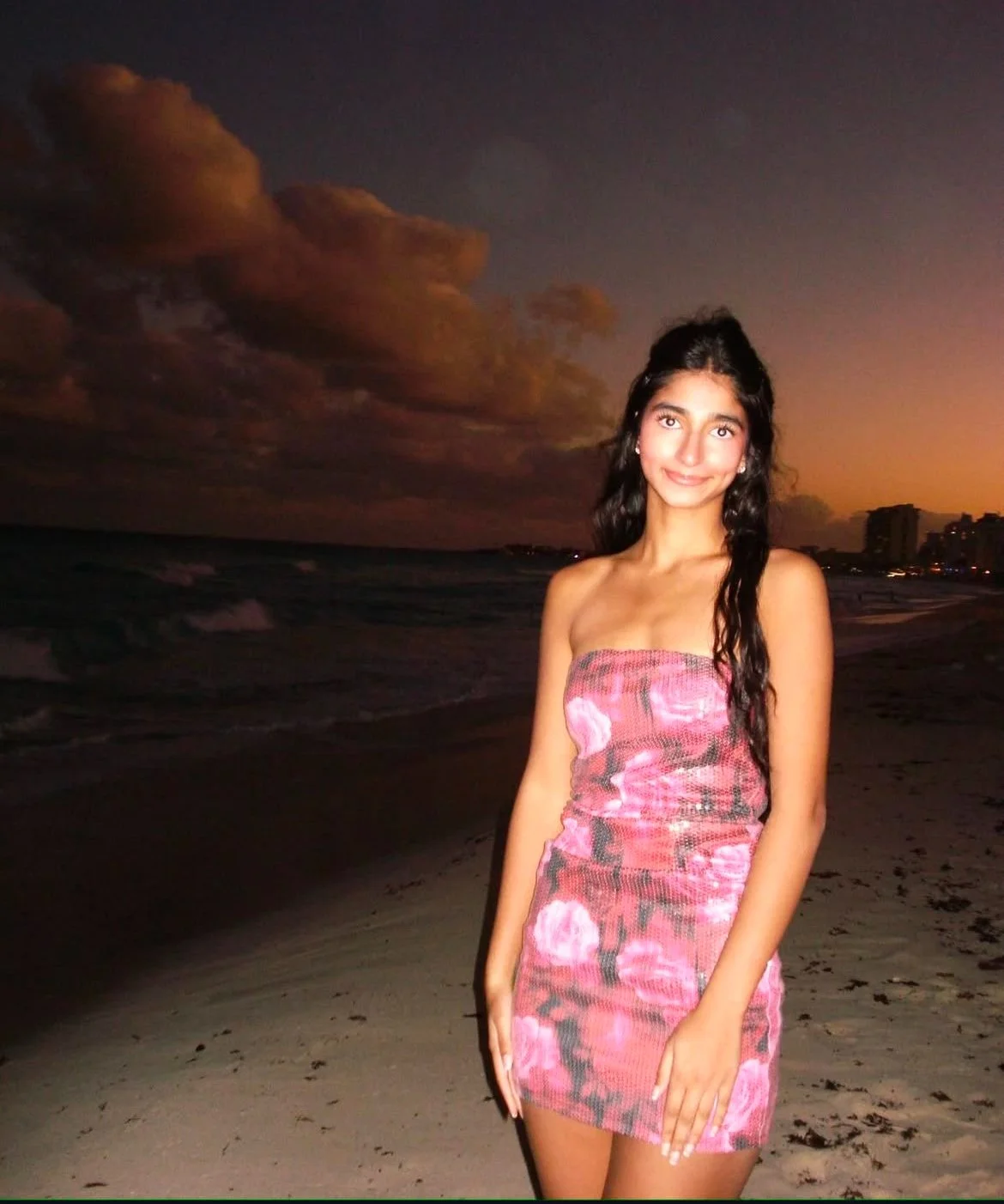 A young woman in a pink floral dress standing on a beach during sunset, with clouds and distant city buildings in the background.
