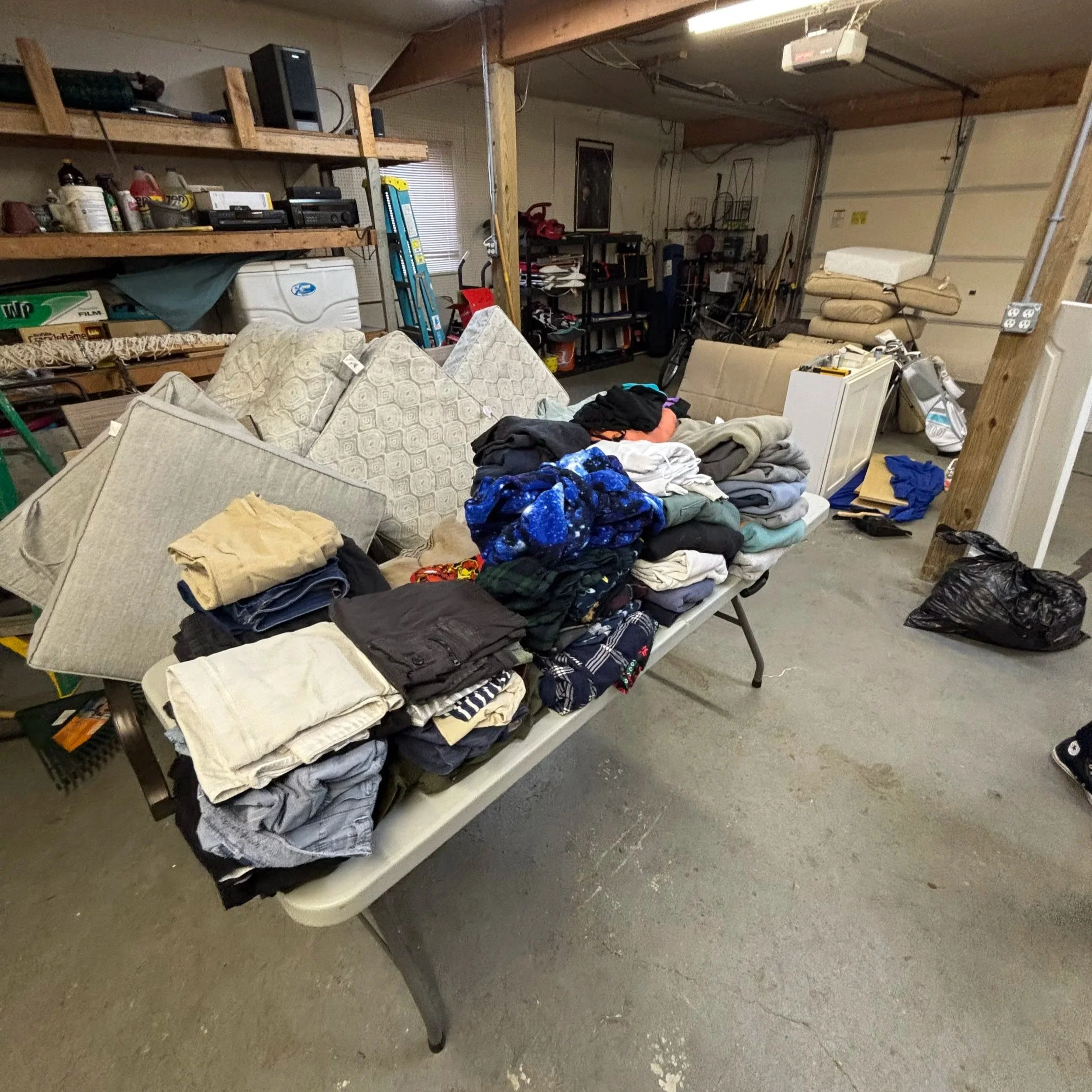 A cluttered garage with shelves filled with bottles, boxes, and electronics. A table with folded clothes and blankets, and various tools and items scattered around.